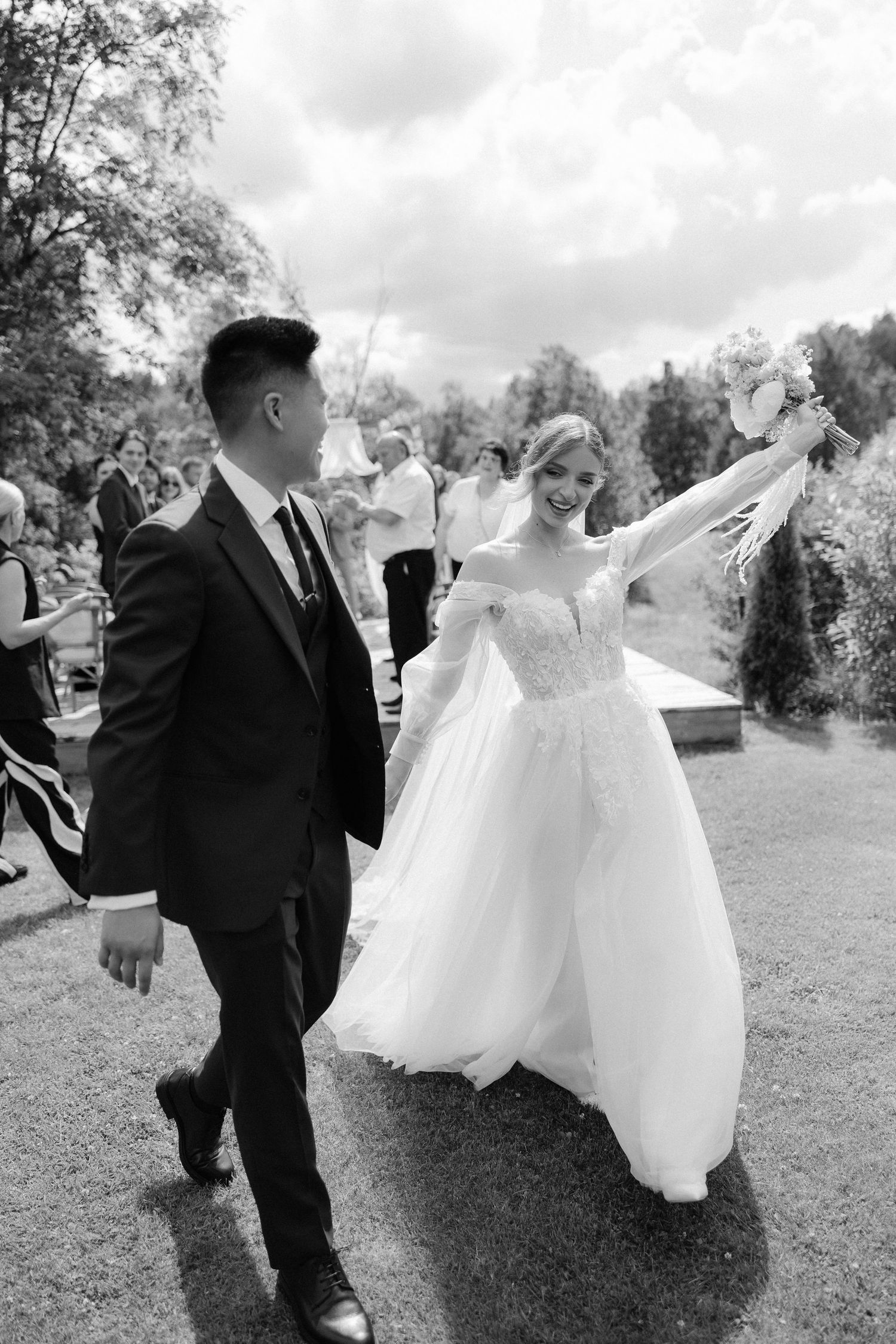 Black and white photo of a joyful bride raising bouquet while walking with groom outdoors at wedding celebration.