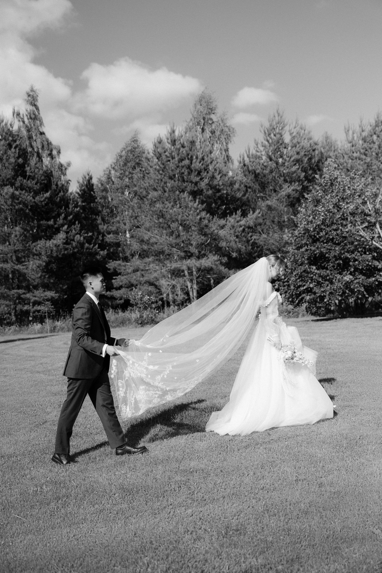 Black and white photo of groom holding bride's flowing lace veil as they walk across lawn.