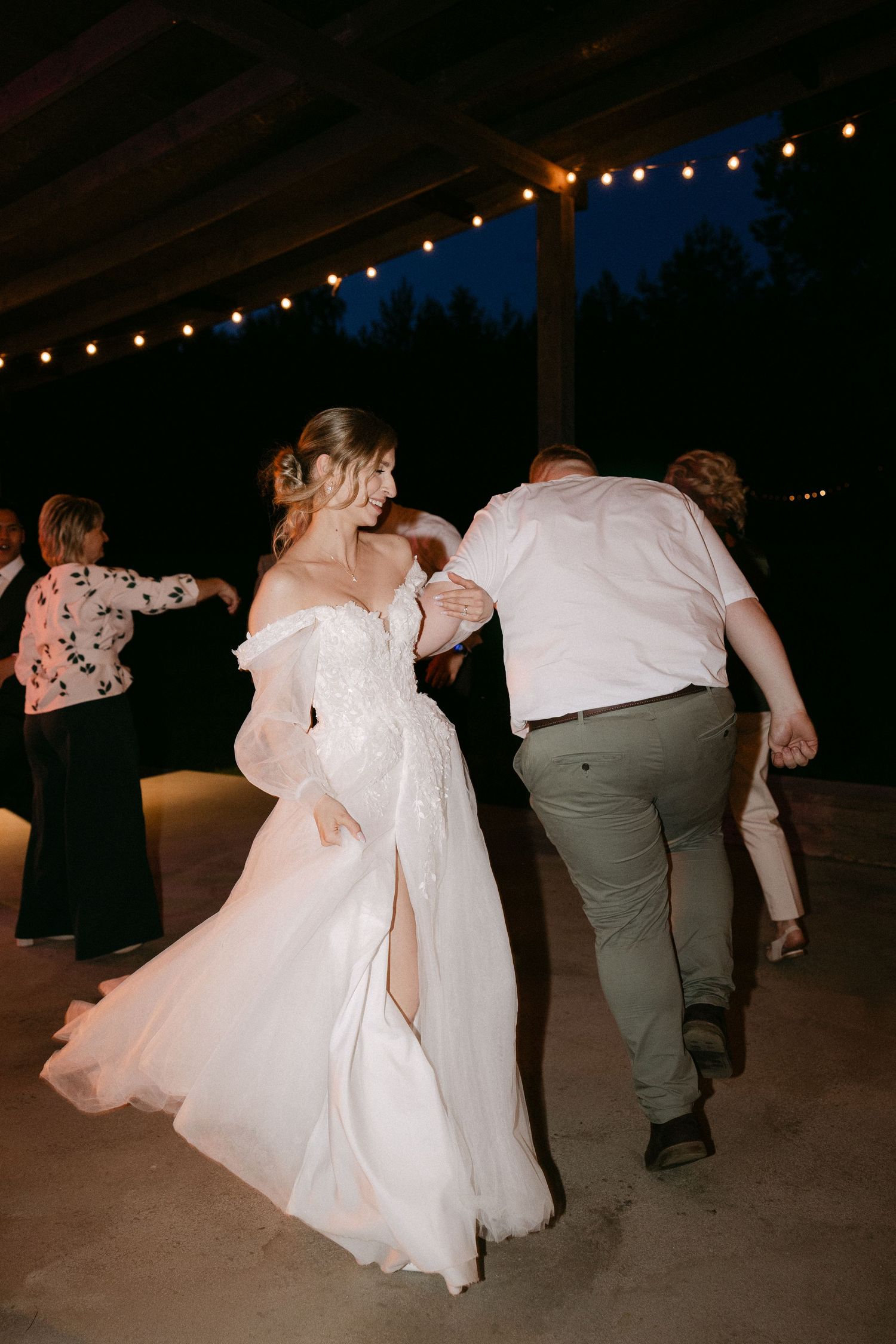 Bride in an off-shoulder lace wedding gown dancing joyfully with a guest at an outdoor evening reception.