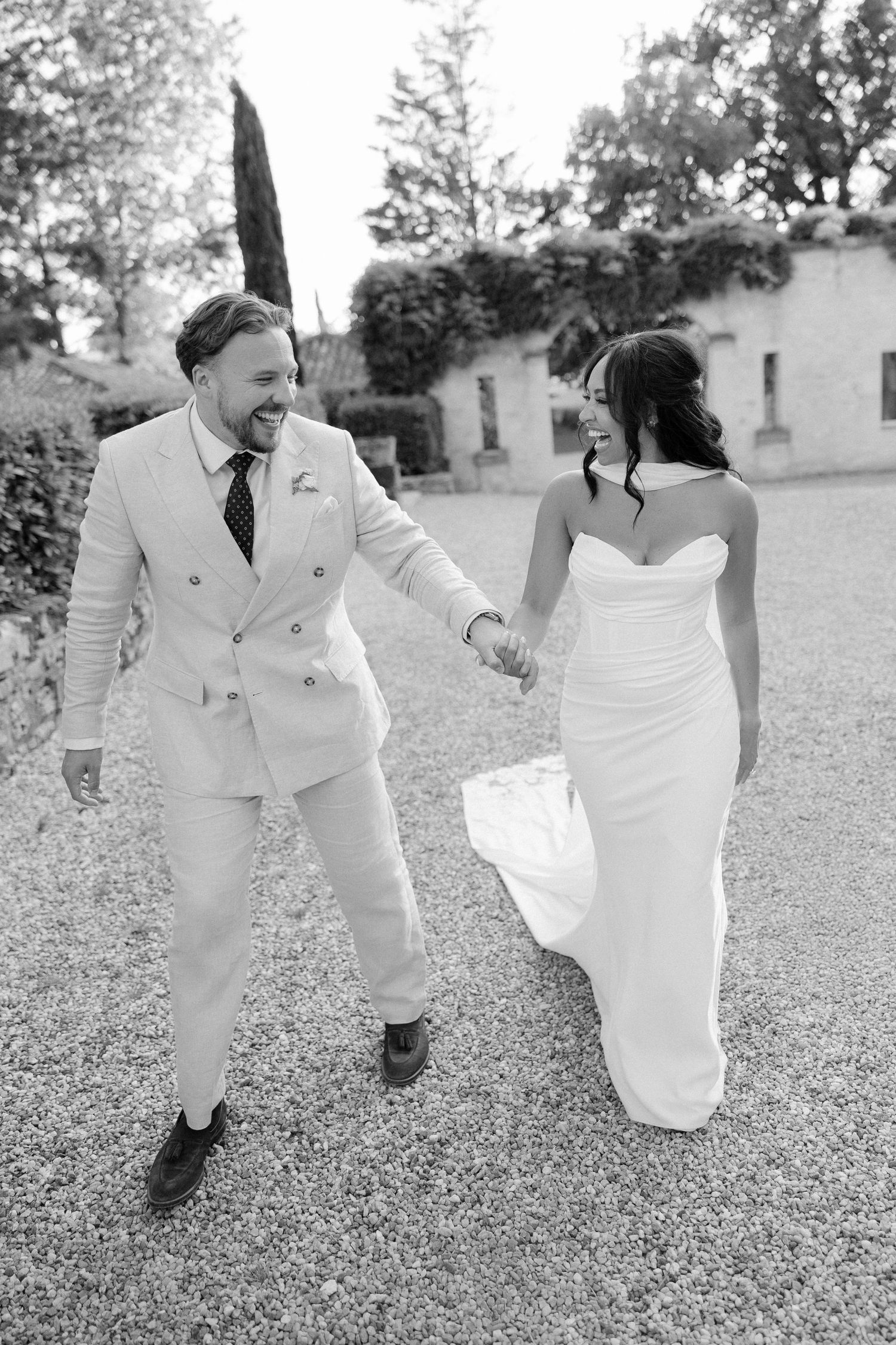 Black and white photo of laughing bride and groom holding hands while walking on a gravel path.