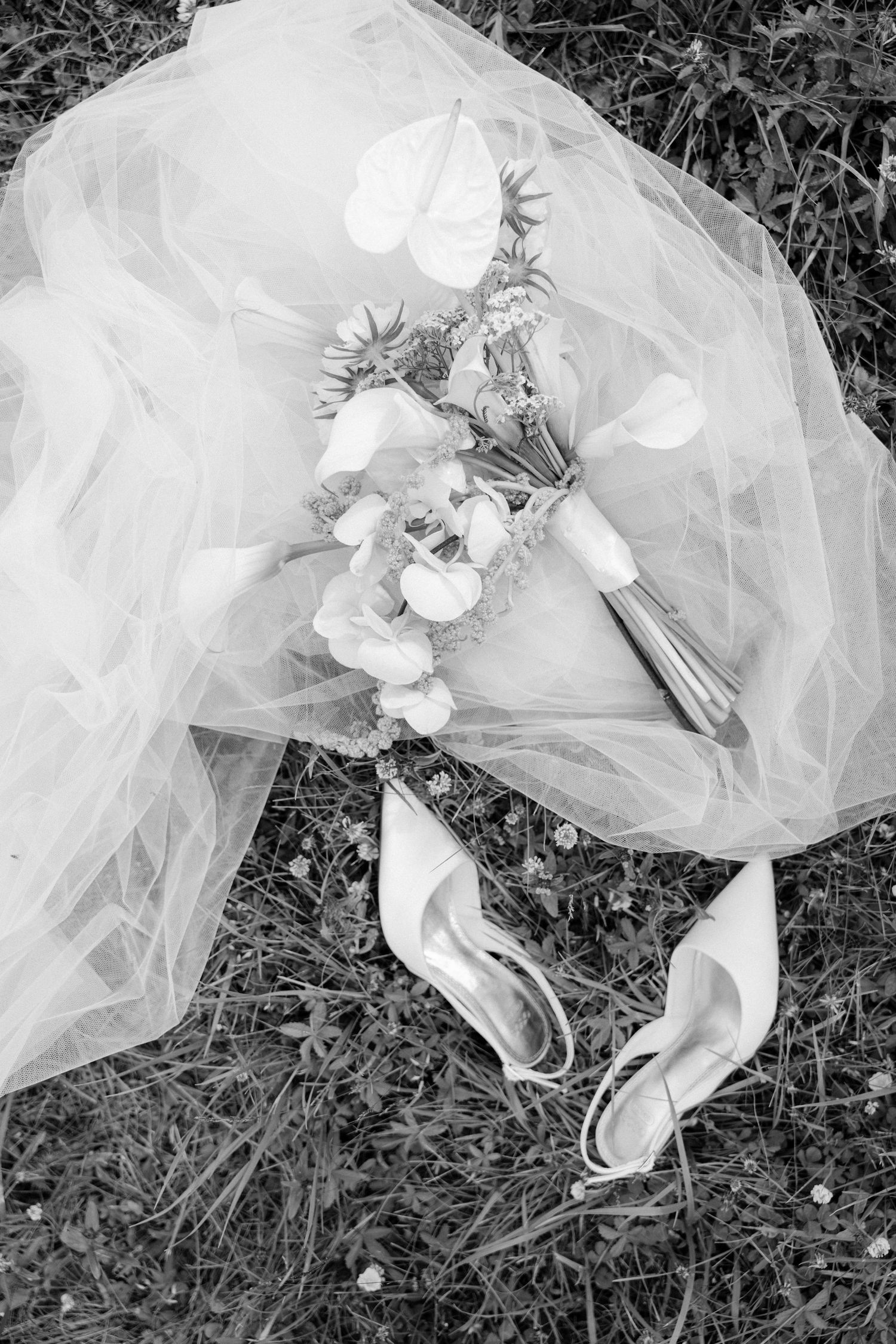 Black and white flat lay of a bridal bouquet, veil, and heels resting on grass.