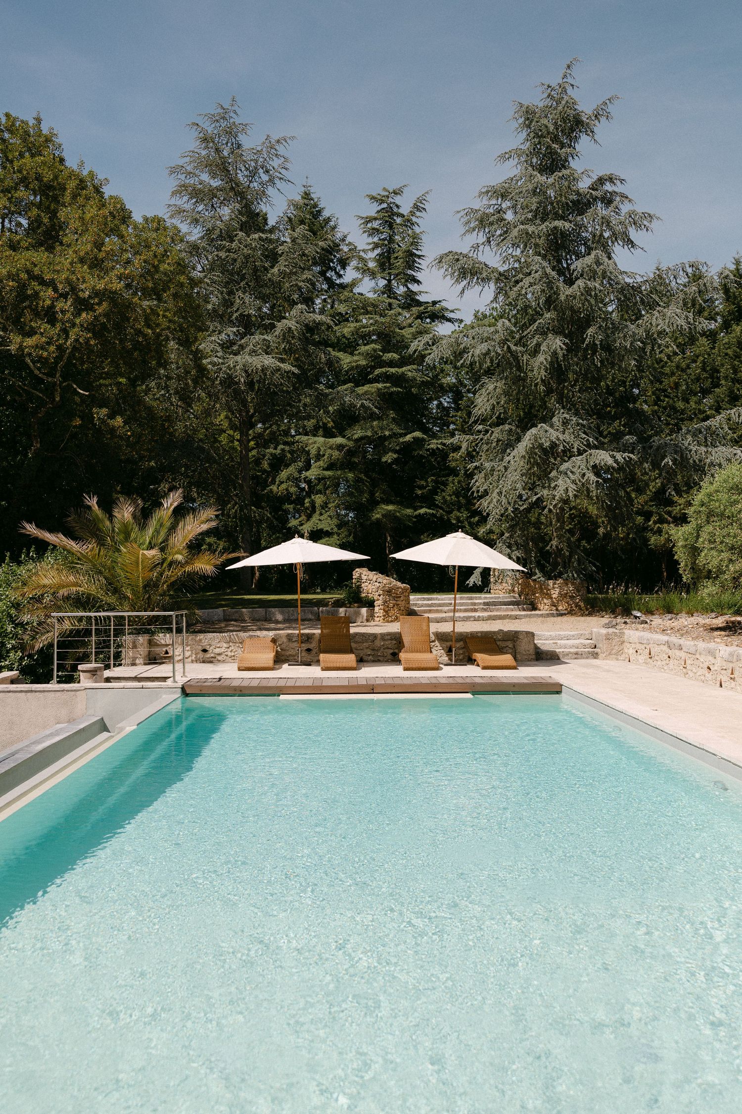 Luxury outdoor swimming pool with lounge chairs and white umbrellas surrounded by lush green trees on a sunny day.