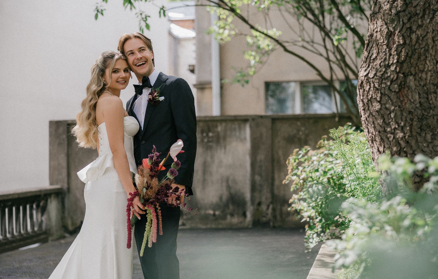 A couple shares romantic moments in their wedding attire while posing outdoors among trees and greenery.