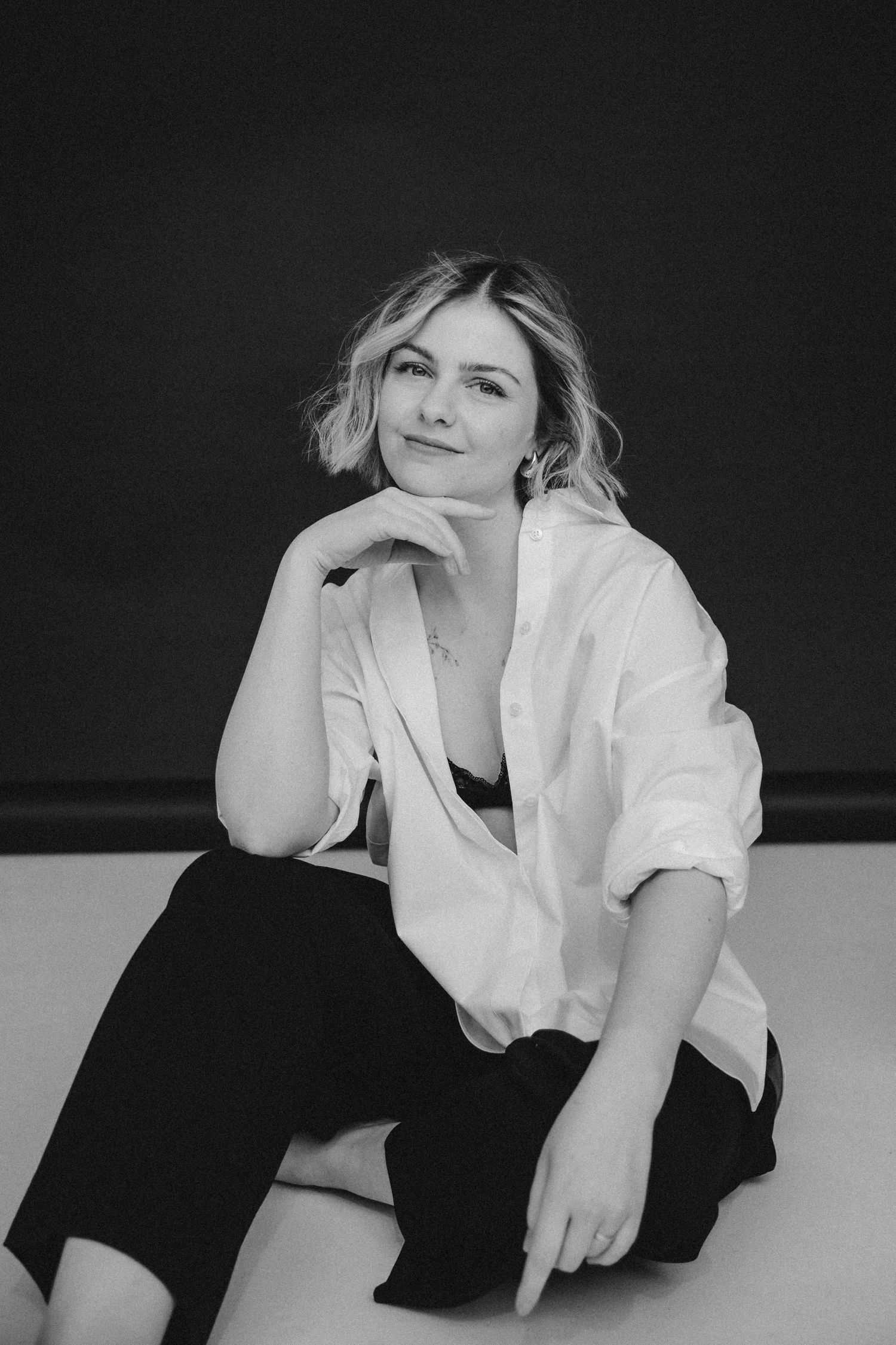 Black and white portrait of a smiling blonde woman in white shirt and black pants sitting gracefully on floor.