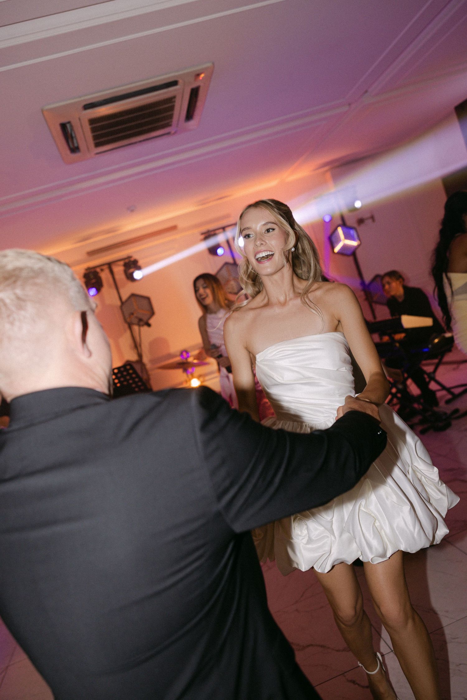 Bride in a short white dress dances joyfully with a guest on a purple-lit reception dance floor.