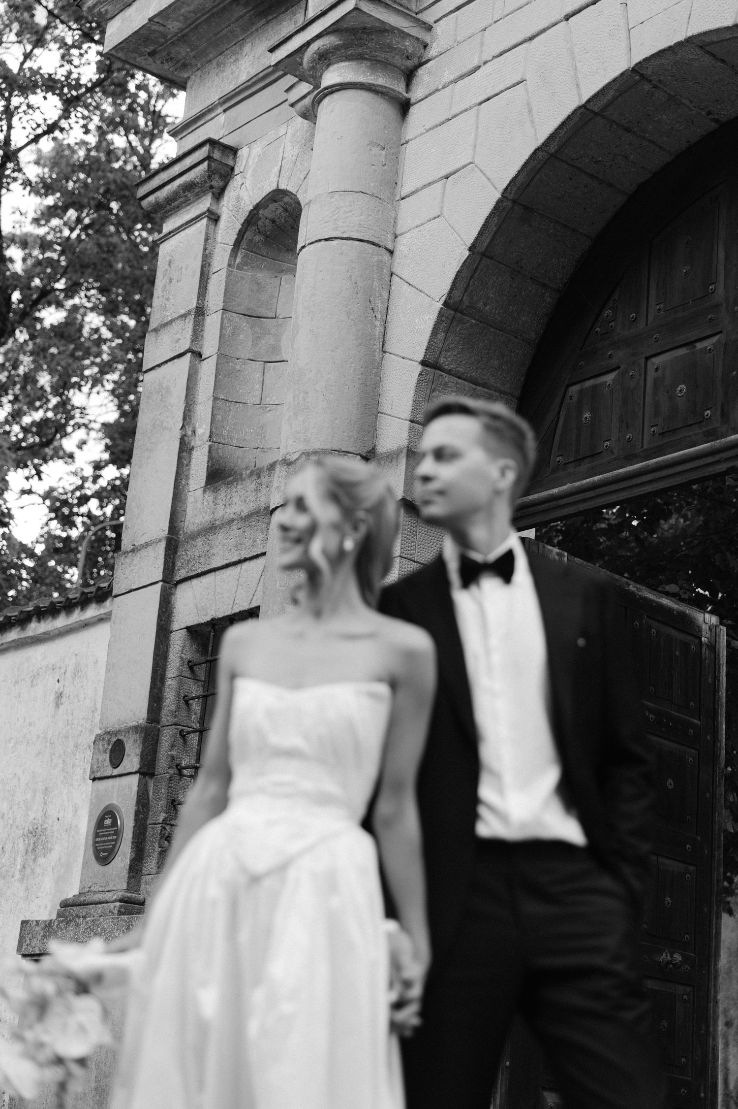 Black and white photo of bride and groom holding hands before ornate stone archway entrance.