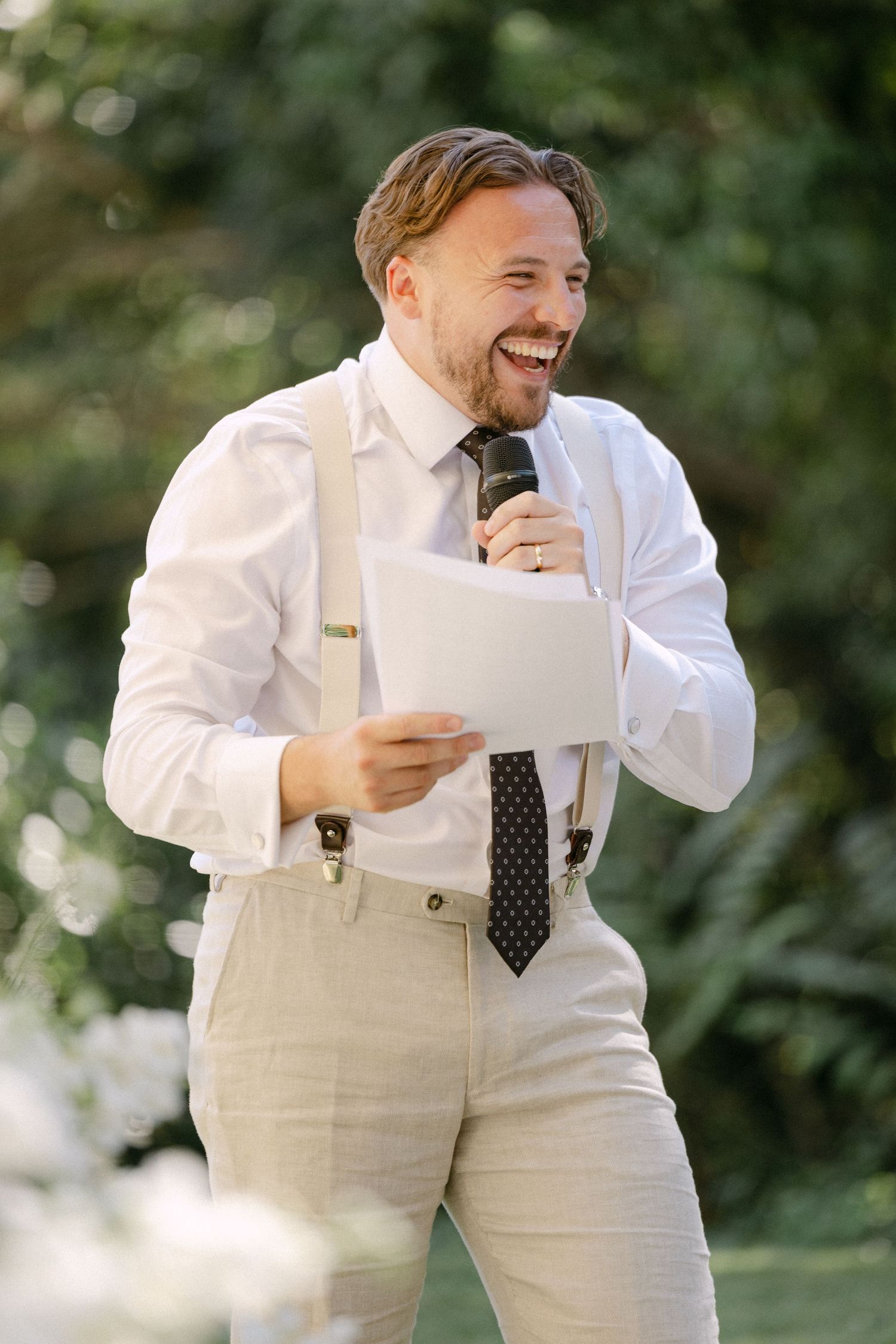 A laughing groomsman in white shirt with suspenders and tie delivers a speech holding a microphone and papers outdoors.