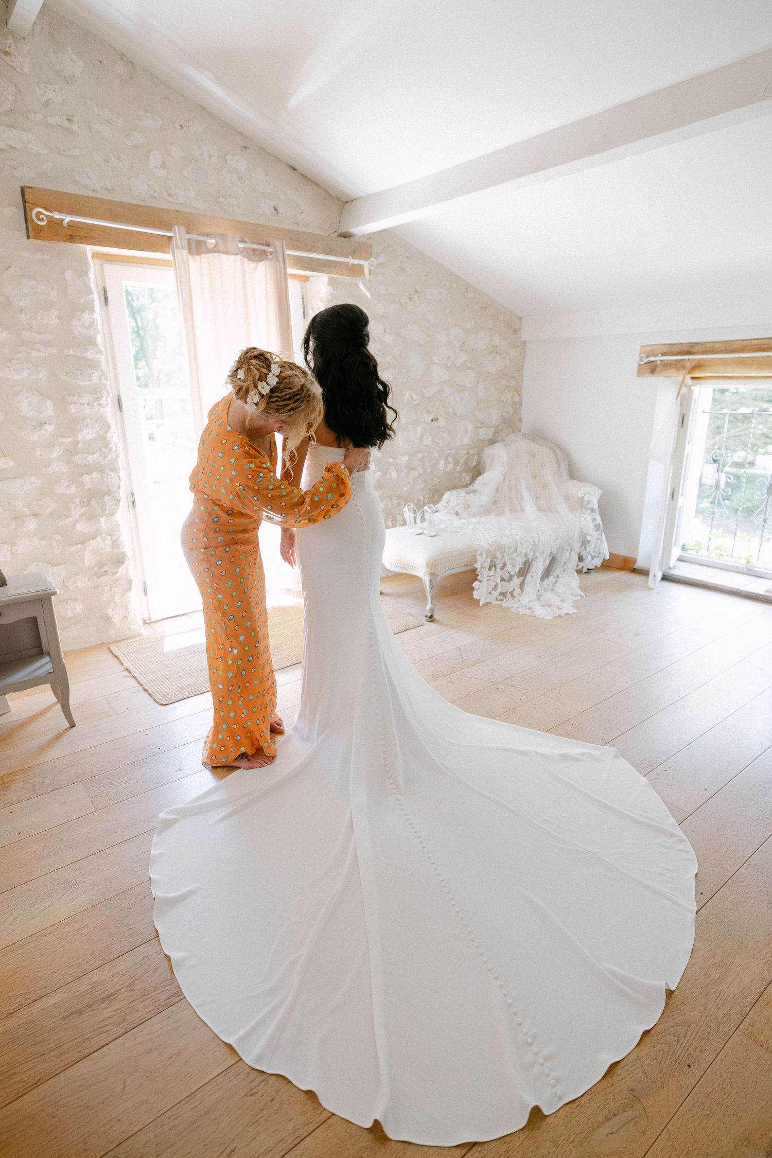 Bridesmaid in orange sequin dress helping bride into fitted white gown with cathedral train in rustic venue.