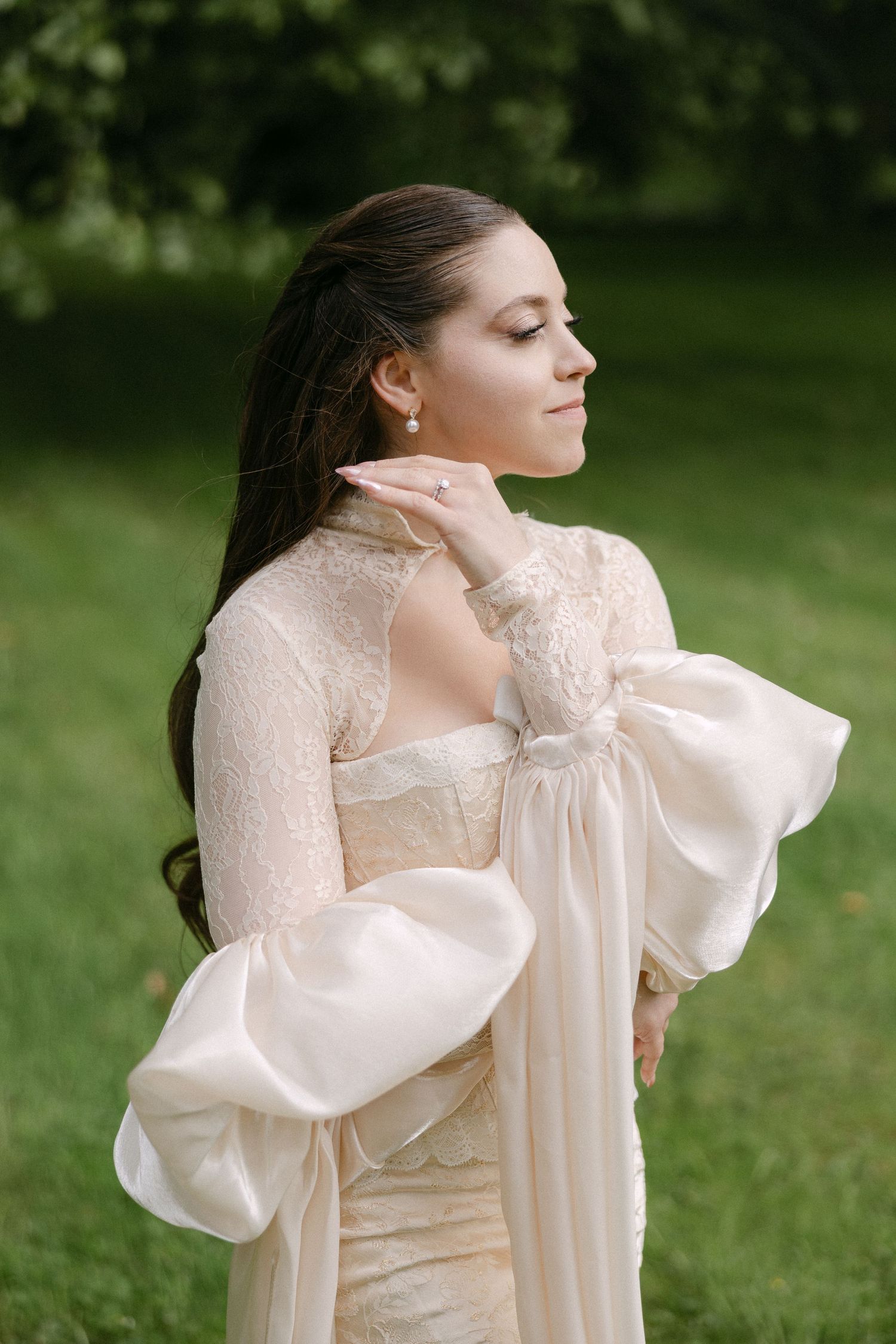 Bride in stunning lace wedding dress with dramatic oversized bow sleeves posing outdoors in green garden.