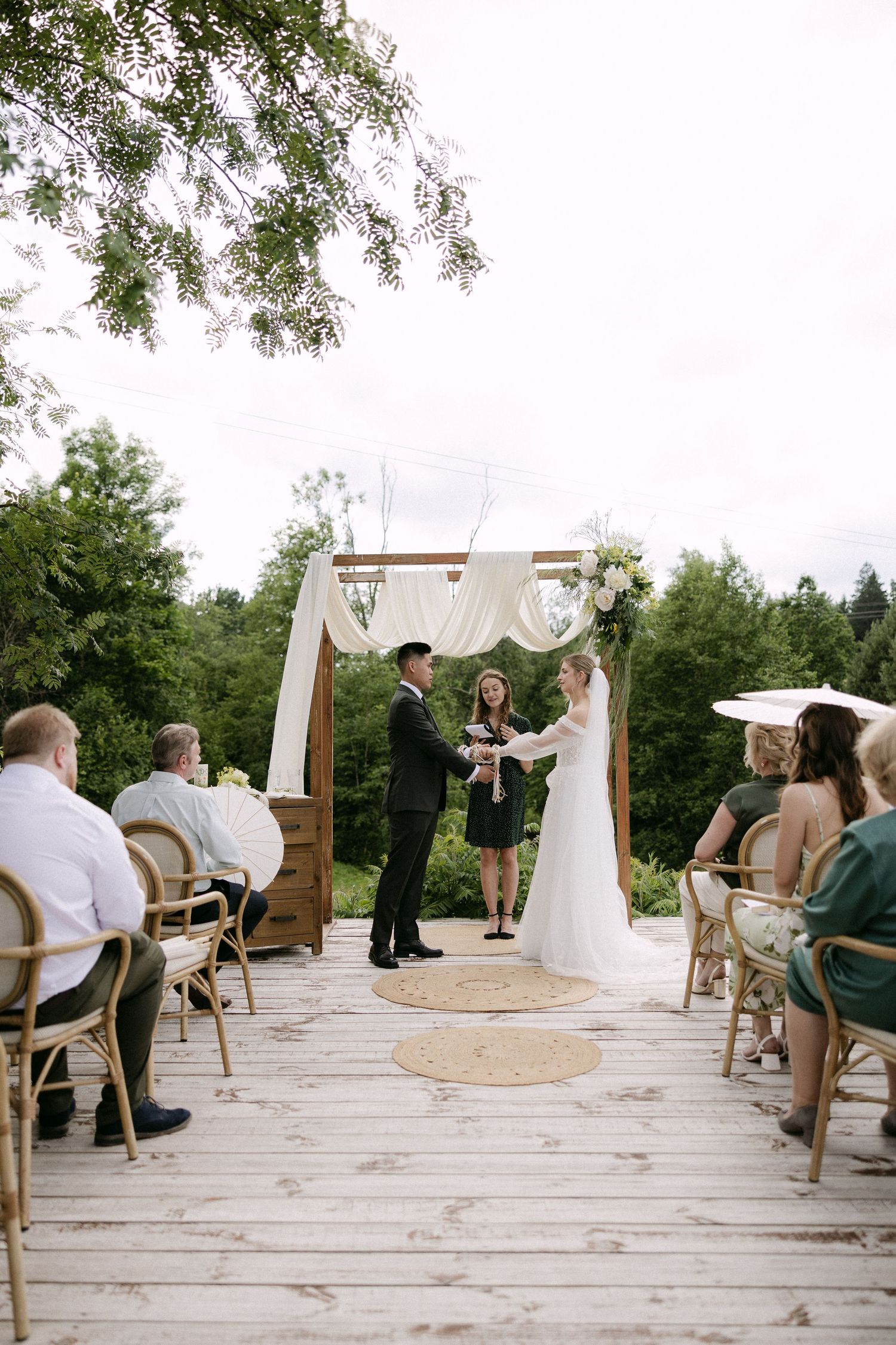 Bride and groom exchanging vows at outdoor wedding ceremony on wooden deck surrounded by greenery.