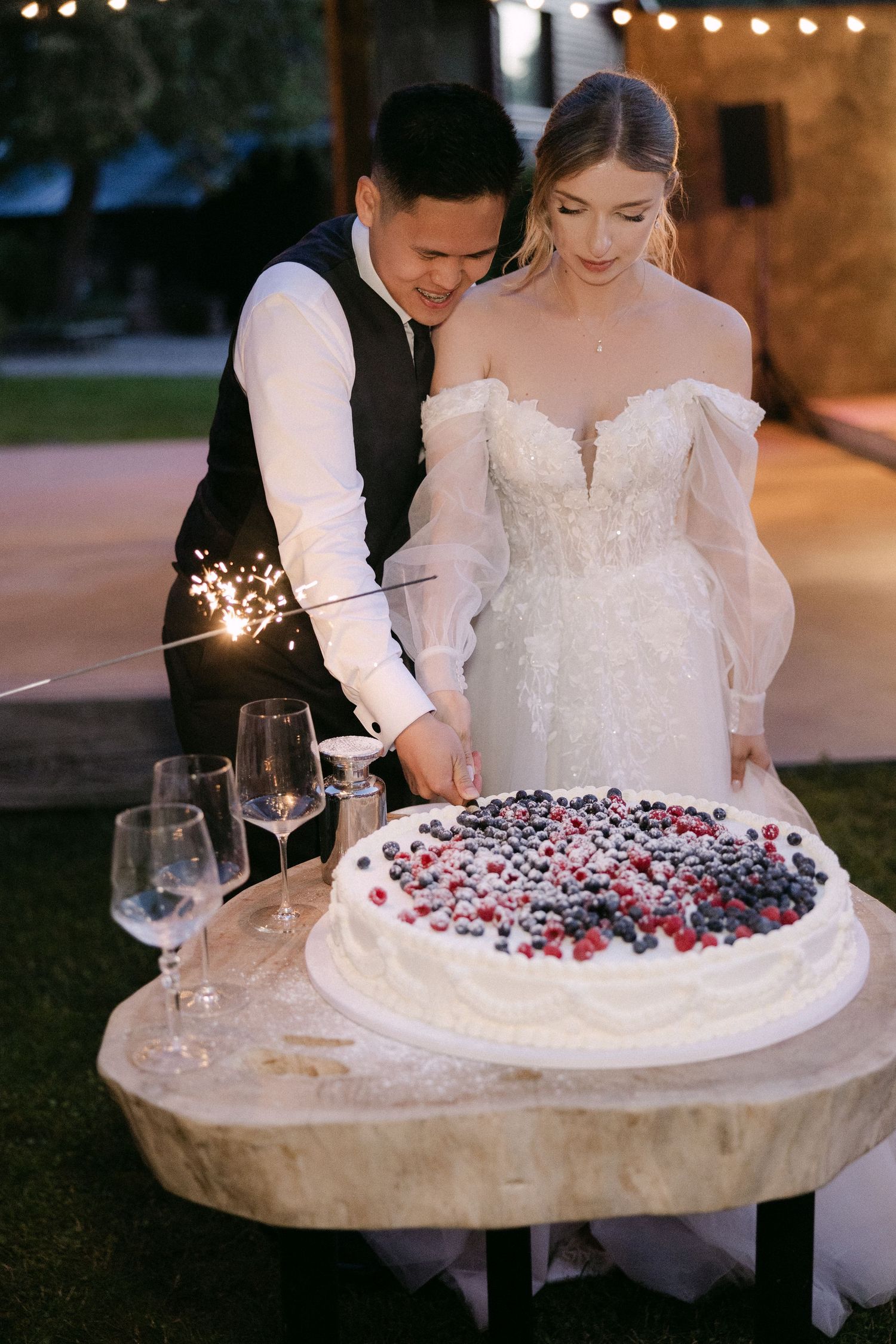 Newlyweds cut white berry-topped wedding cake together with sparklers at reception.