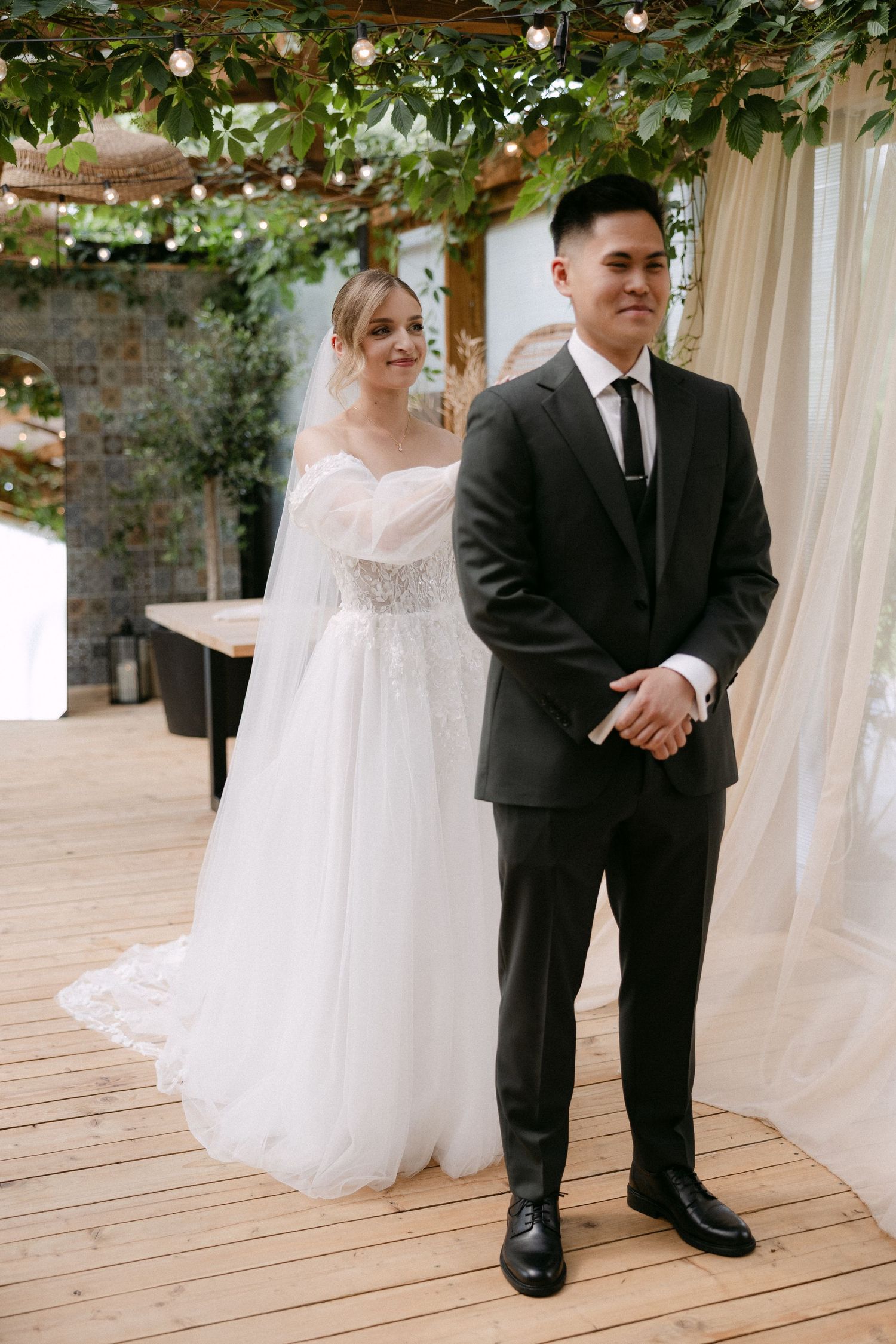 Bride in white lace gown and groom in black suit standing at wedding ceremony altar with greenery overhead.