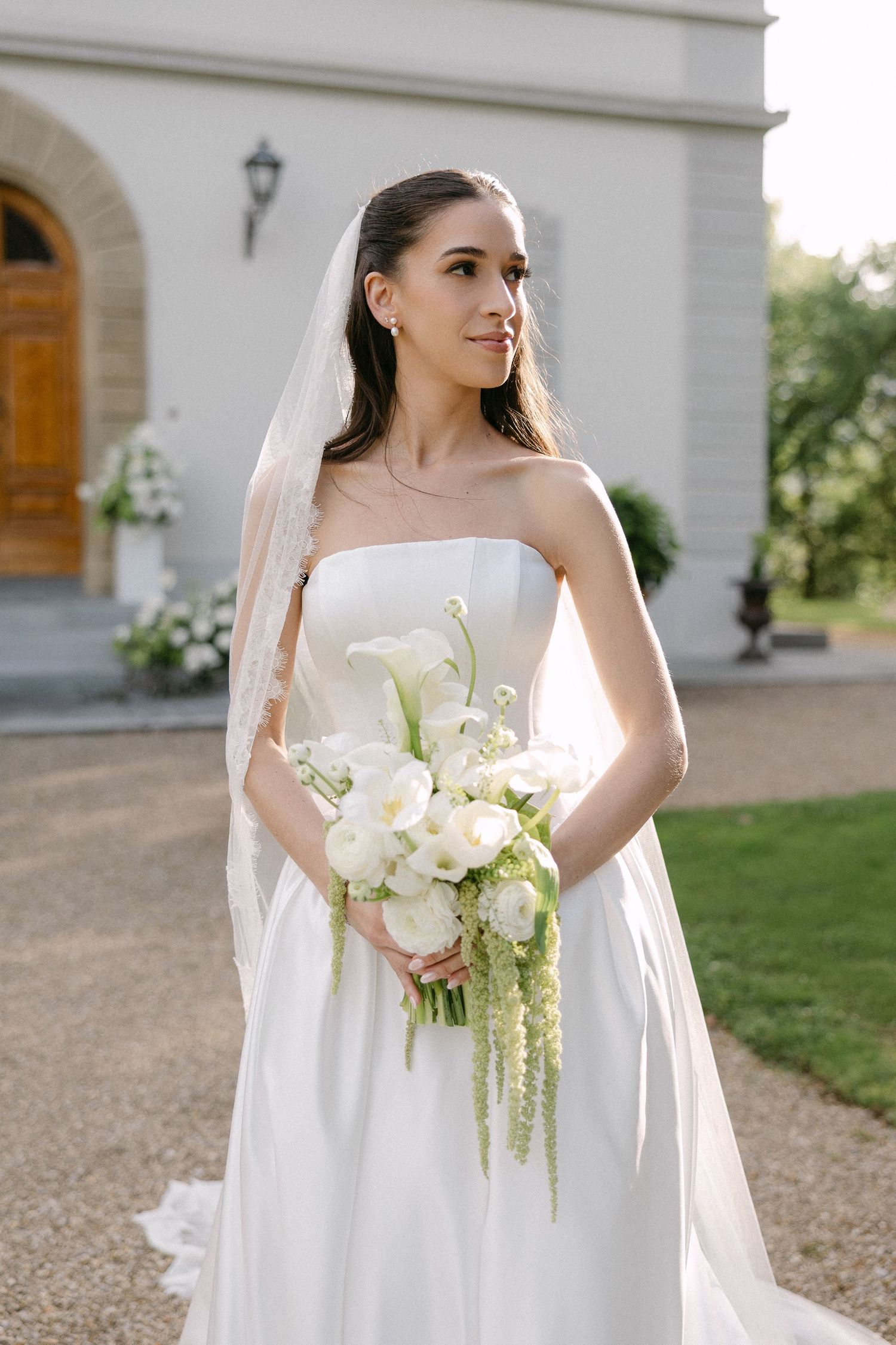 Elegant bride in strapless white gown holds lush white calla lily bouquet while wearing cathedral veil outdoors.