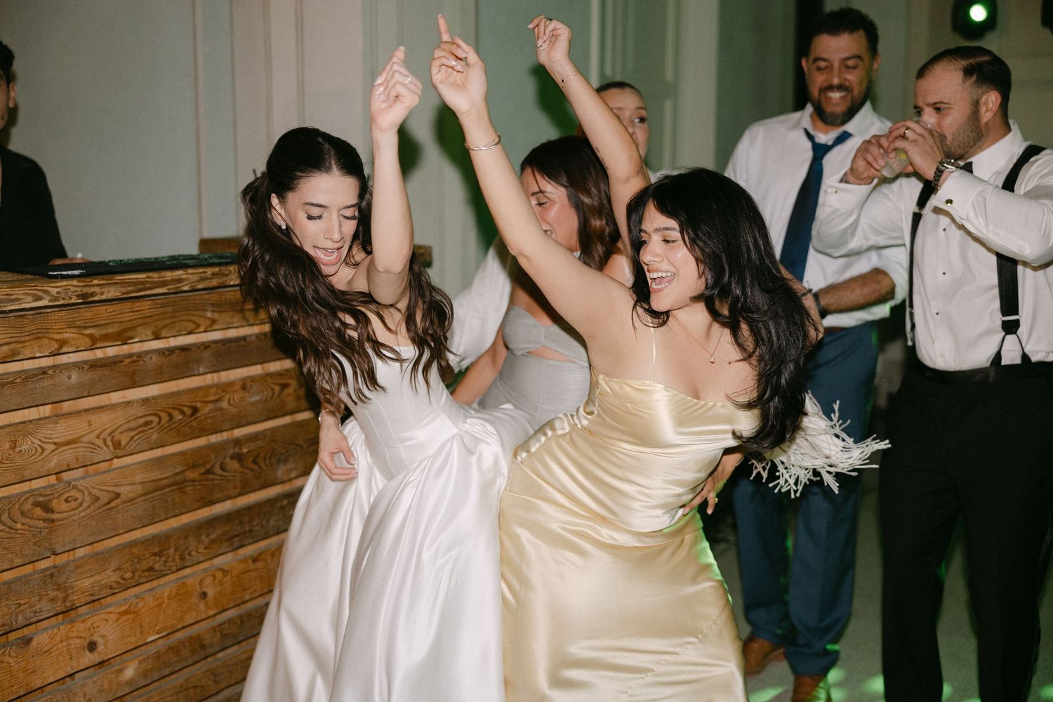 Bride and bridesmaid in gold dress dancing with arms raised joyfully at lively wedding reception.