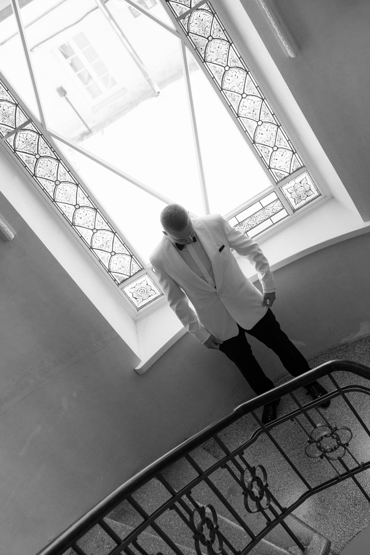 Man in white tuxedo jacket stands on elegant spiral staircase near ornate stained glass windows in black and white photo.