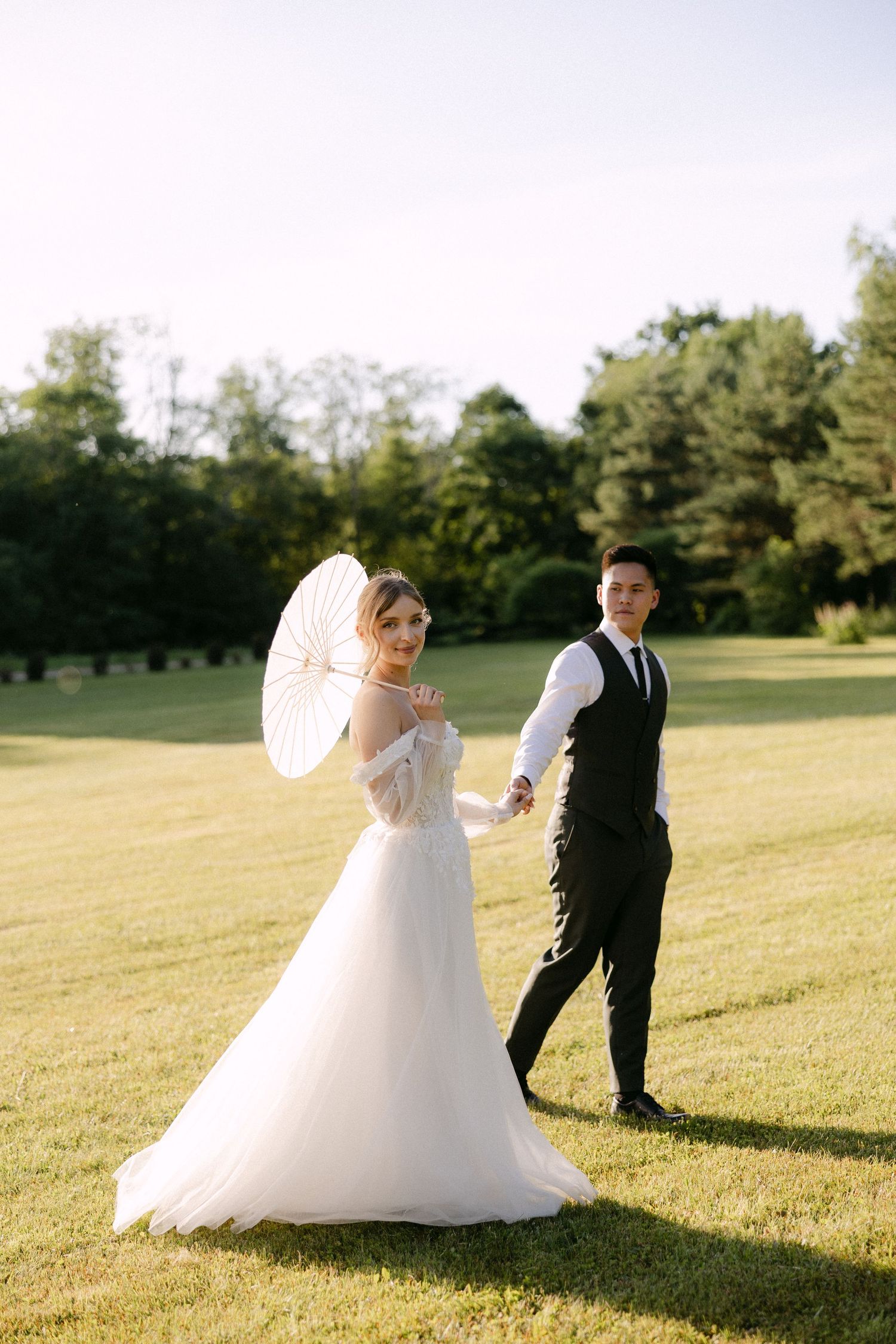 Bride in white gown holding parasol walks hand-in-hand with groom in black suit across lush green lawn.
