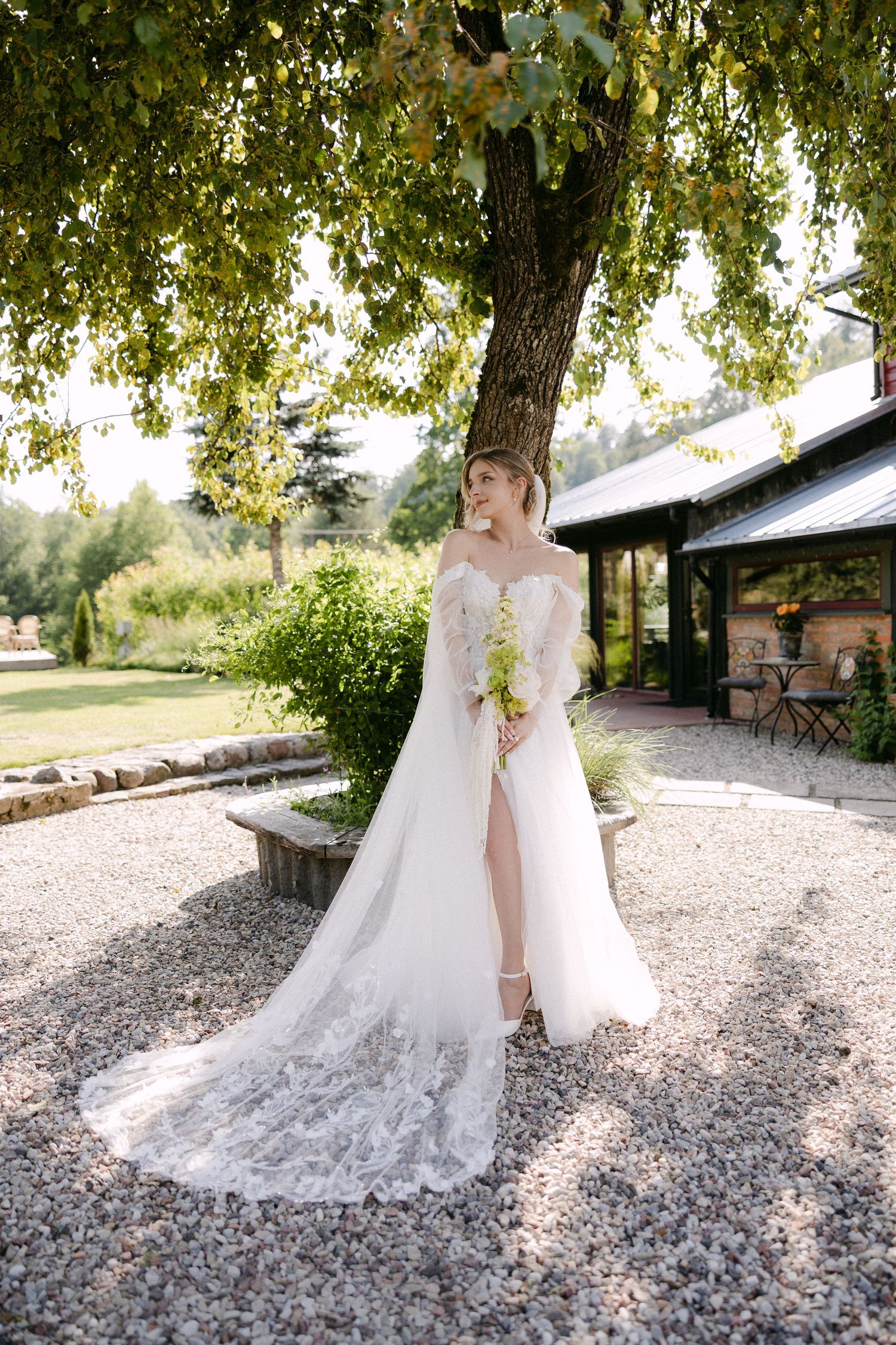 Bride in off-shoulder lace wedding gown with train stands under a tree holding yellow flowers in a garden.