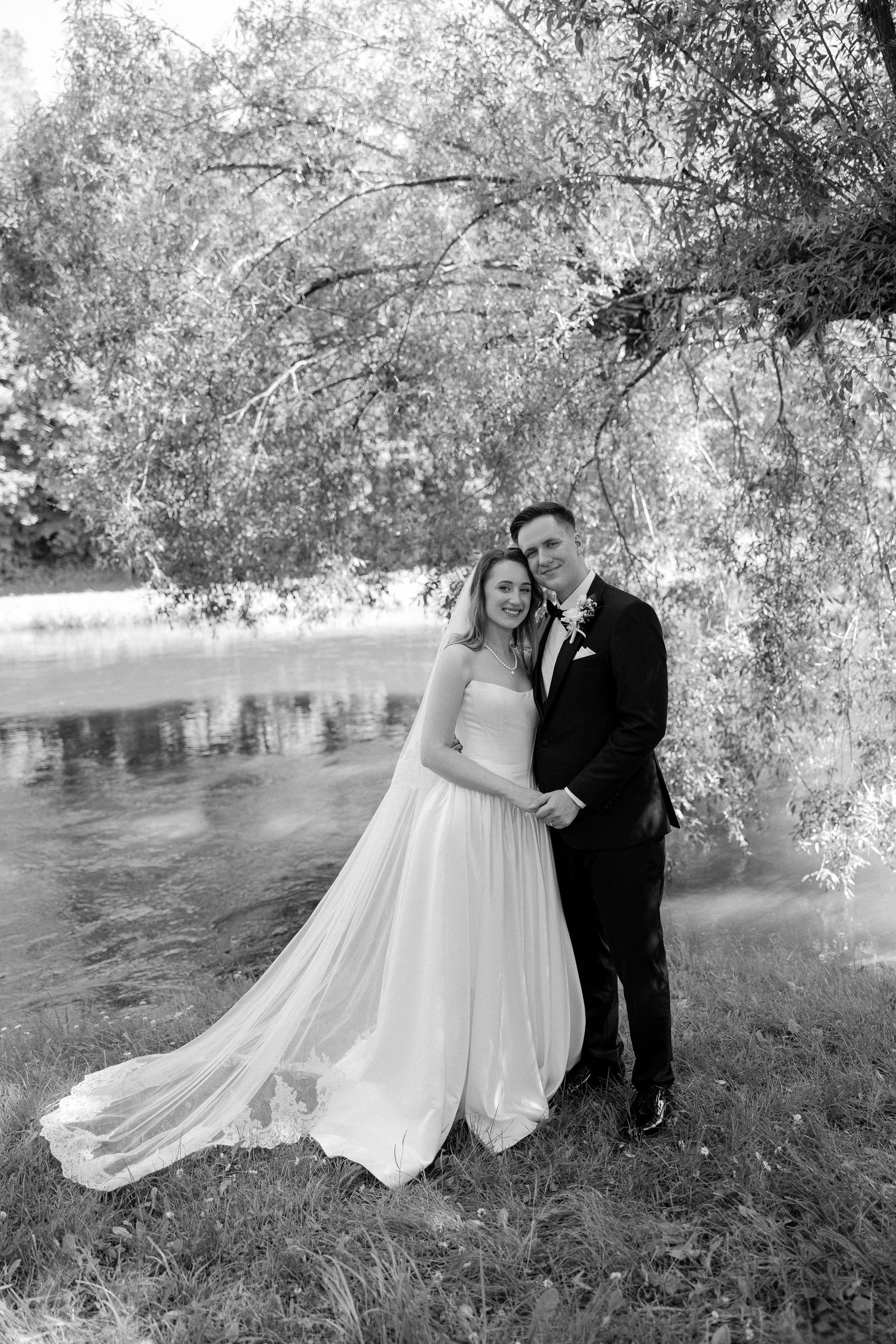 Black and white photo of bride and groom embracing by a serene lake under weeping willow trees.