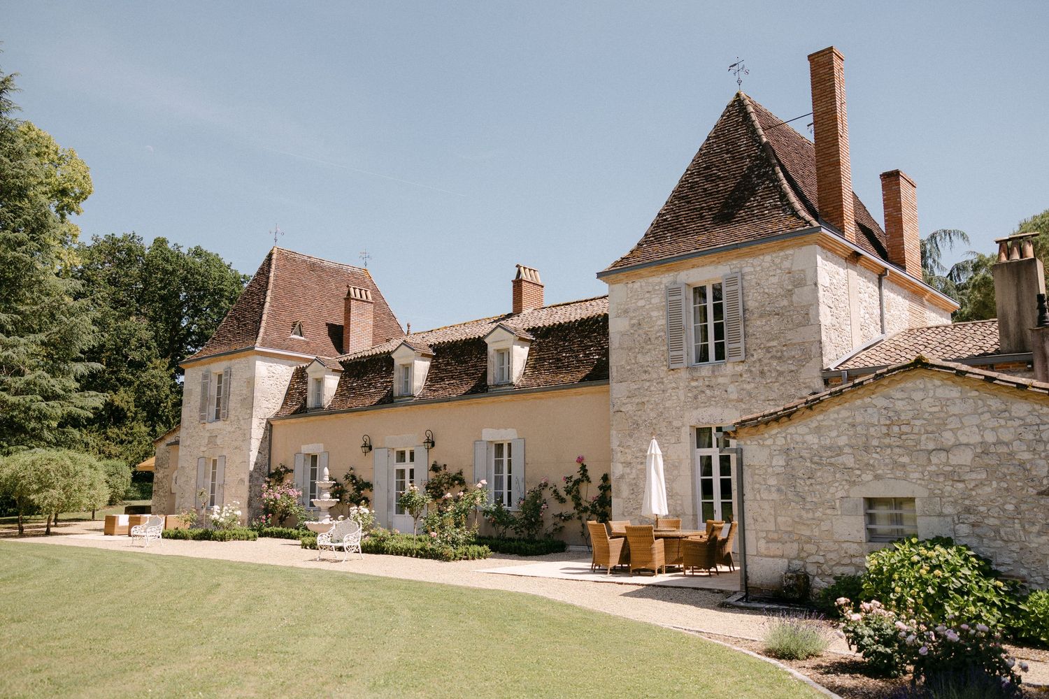 Elegant French chateau wedding venue with stone facade, manicured lawns, and terracotta roof tiles under a clear blue sky.
