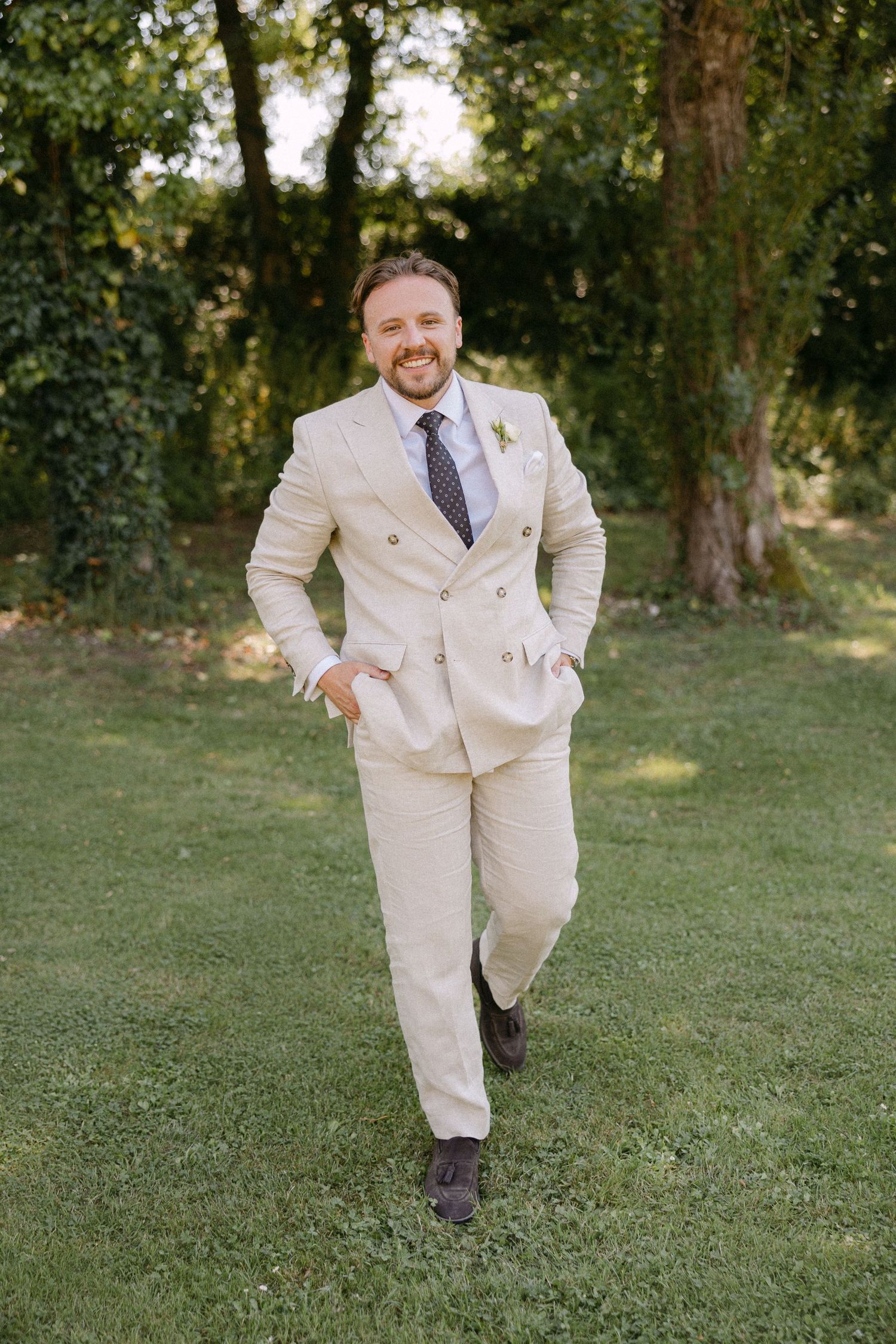 Smiling groom in cream double-breasted suit with dark tie walking on lush green lawn.