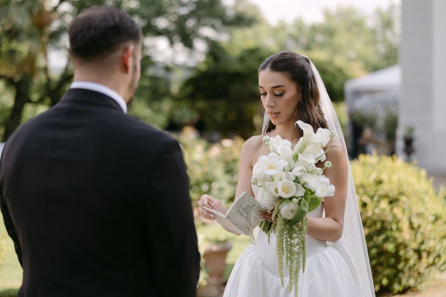 Bride in white gown holding elegant bouquet of white flowers during outdoor wedding ceremony.