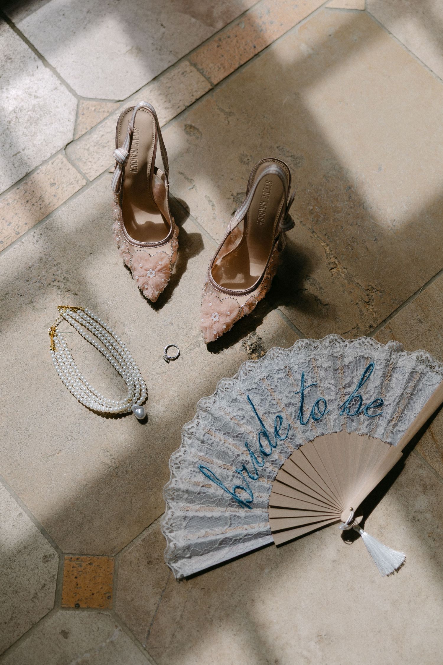 Bridal accessories flat lay featuring lace heels, pearl necklace, ring, and a bride-to-be fan on stone floor.