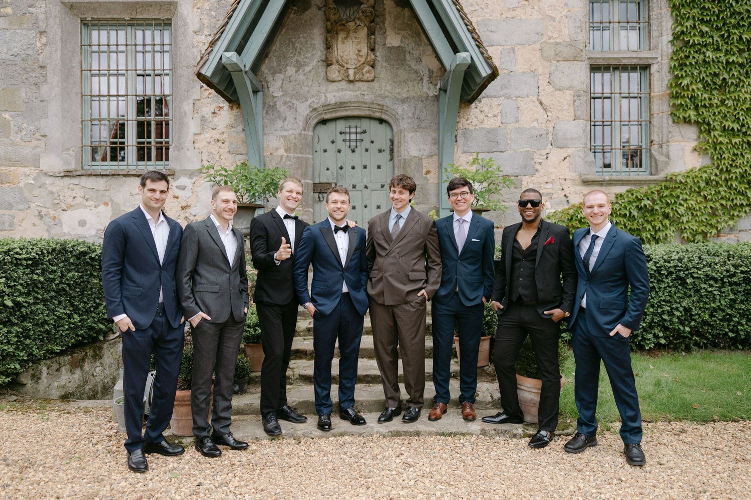Group of eight groomsmen posing in suits outside a historic stone manor house entrance.