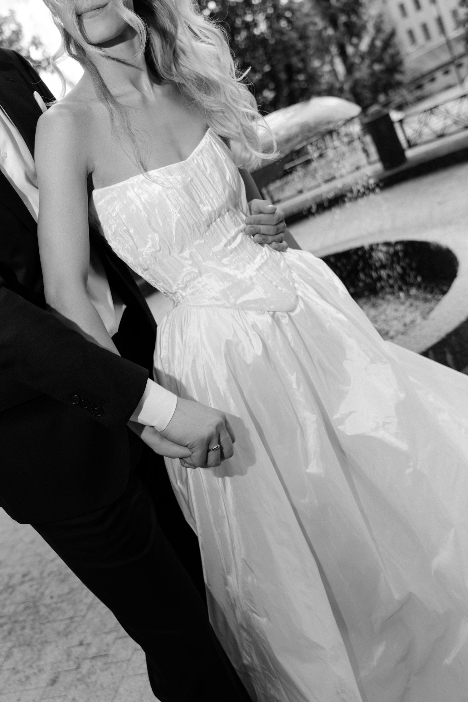 Black and white photo of a bride in a strapless gown holding hands with a groom in a suit at an outdoor wedding.