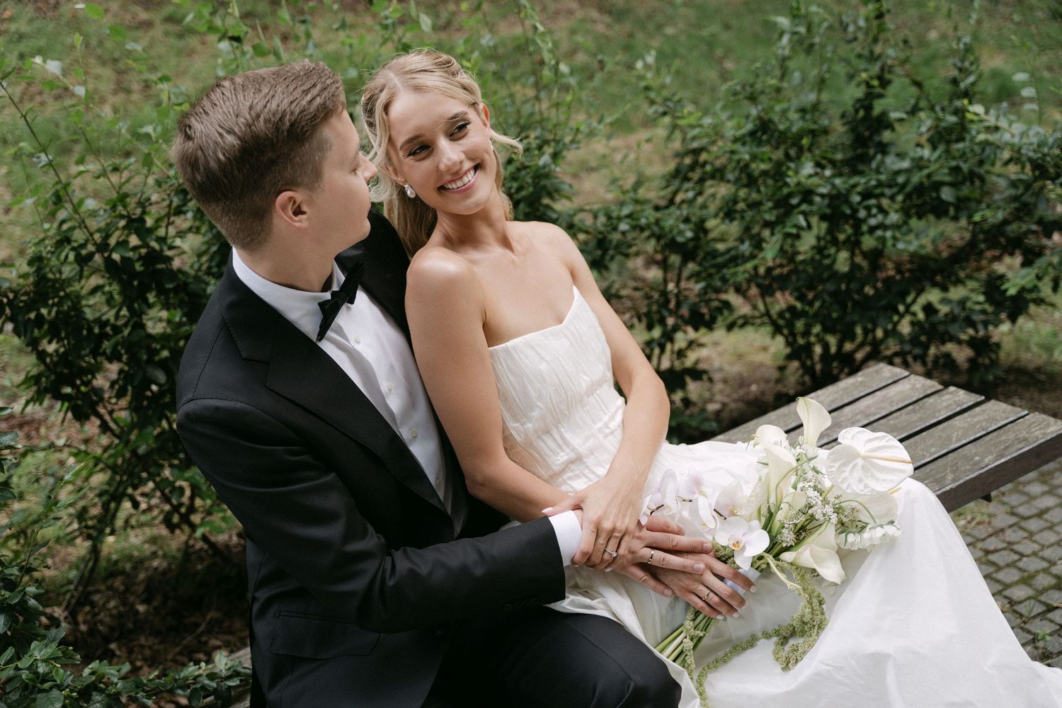 Bride and groom sit together on a bench surrounded by lush greenery, sharing a romantic moment.