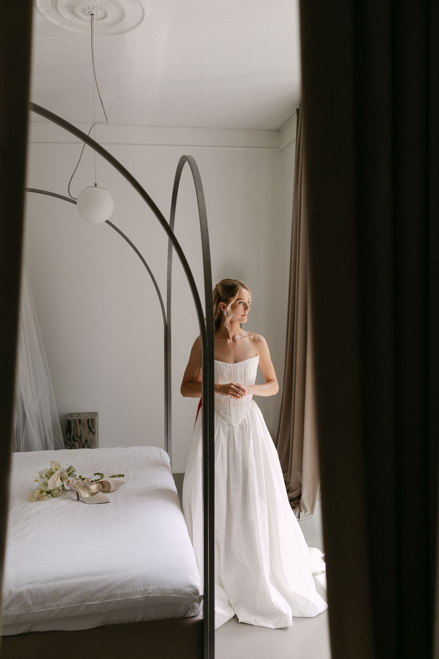 Bride in white strapless wedding gown standing in elegant bedroom with modern arc lamp and bouquet on bed.