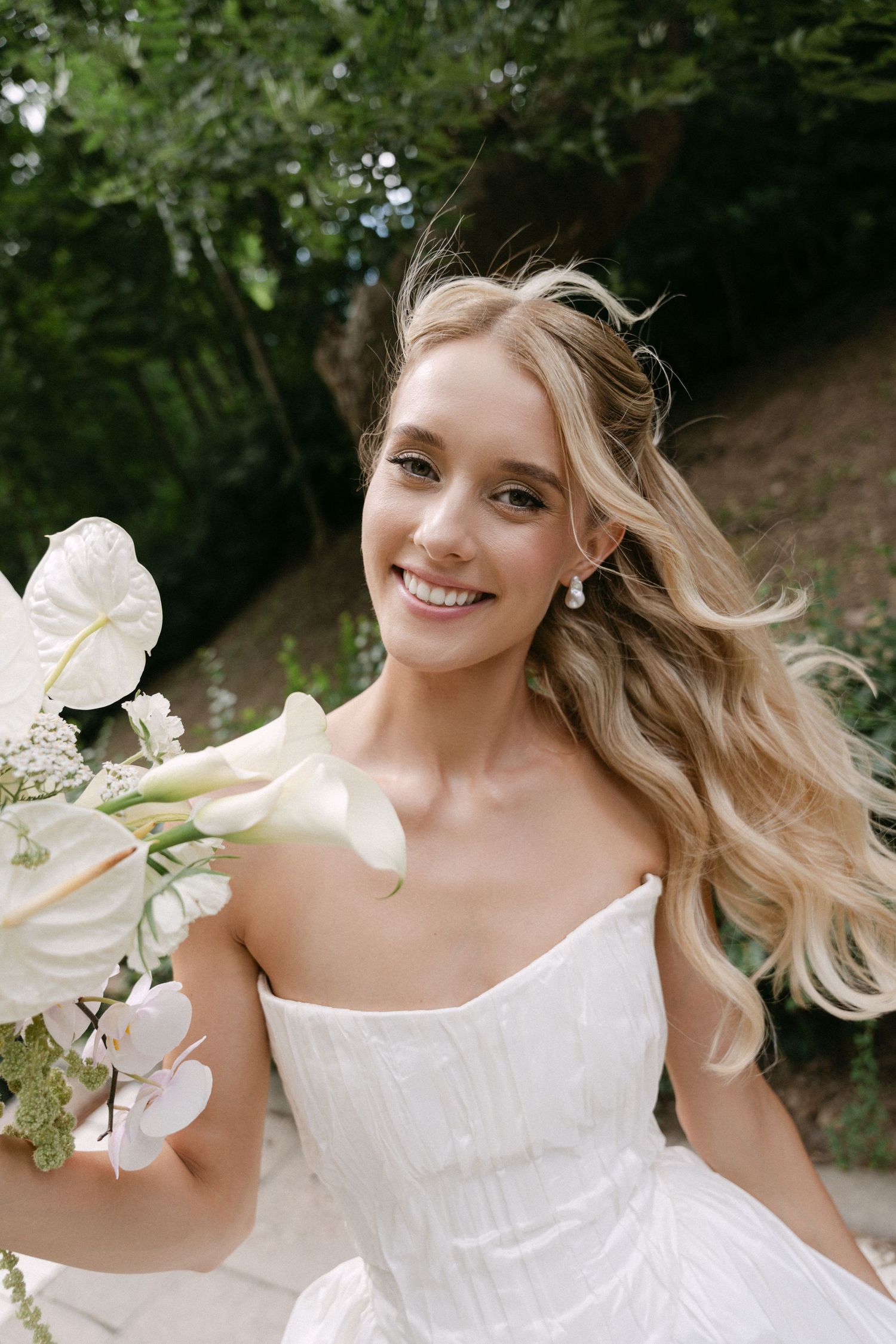 Smiling blonde bride in a strapless white gown holding a white floral bouquet outdoors in a garden.