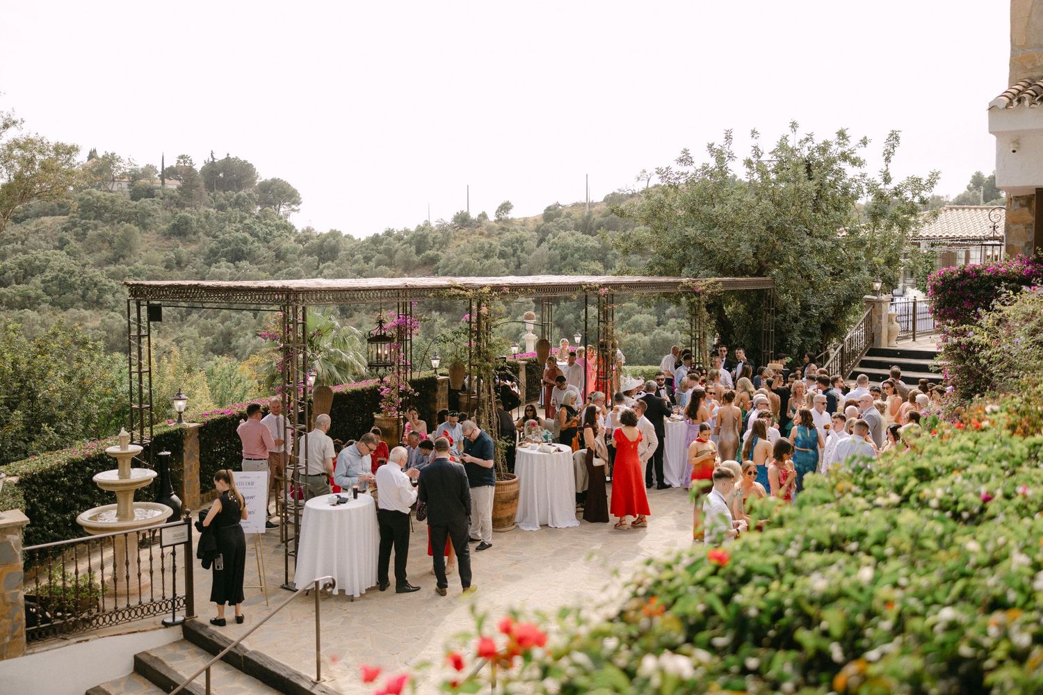 Elegant outdoor wedding reception on a terrace surrounded by lush greenery with guests mingling around white tables.