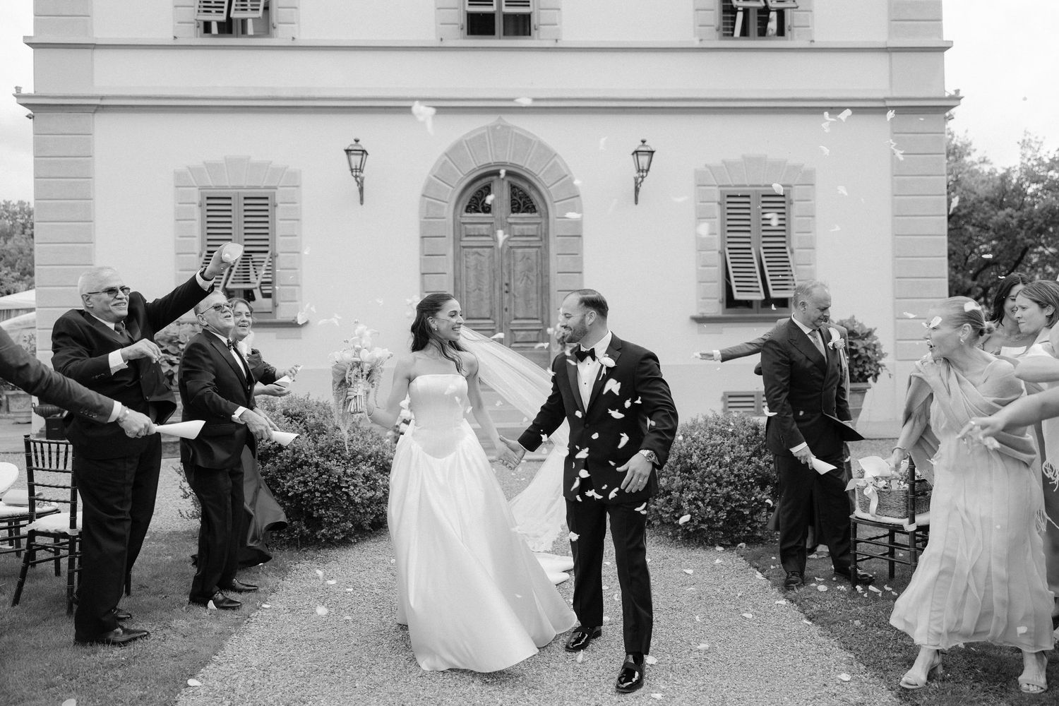 Black and white photo of a bride and groom celebrating with guests tossing confetti outside an elegant villa.