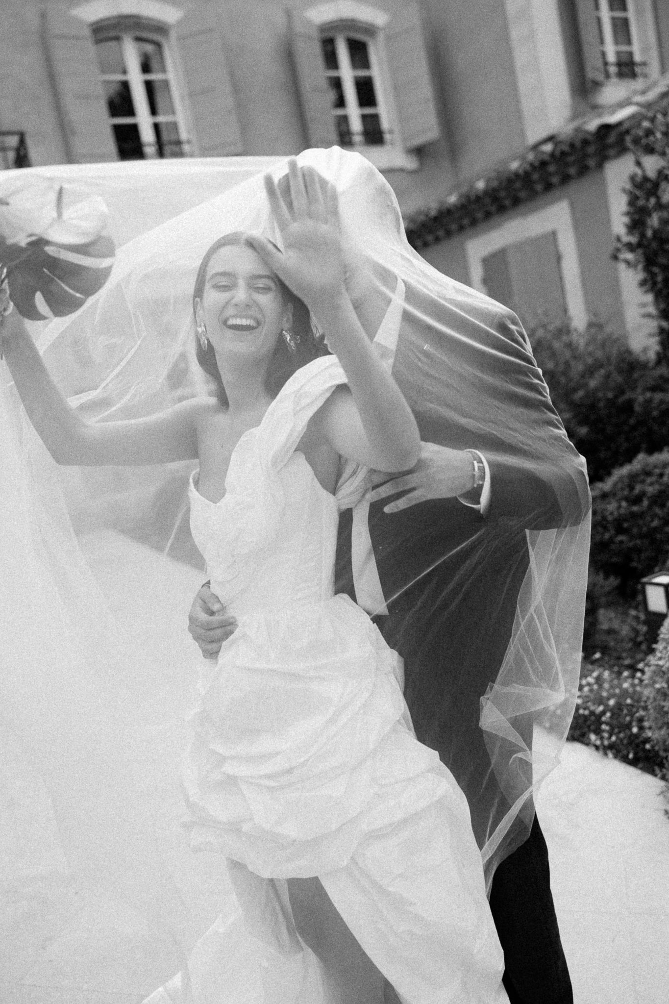 Black and white photo of a laughing bride in a white gown and groom dancing under her flowing veil outside a French château.