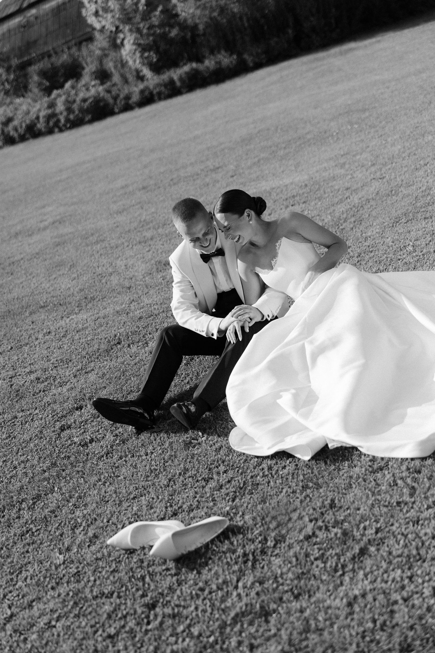 Black and white photo of bride and groom sitting together on grass laughing with shoes nearby.