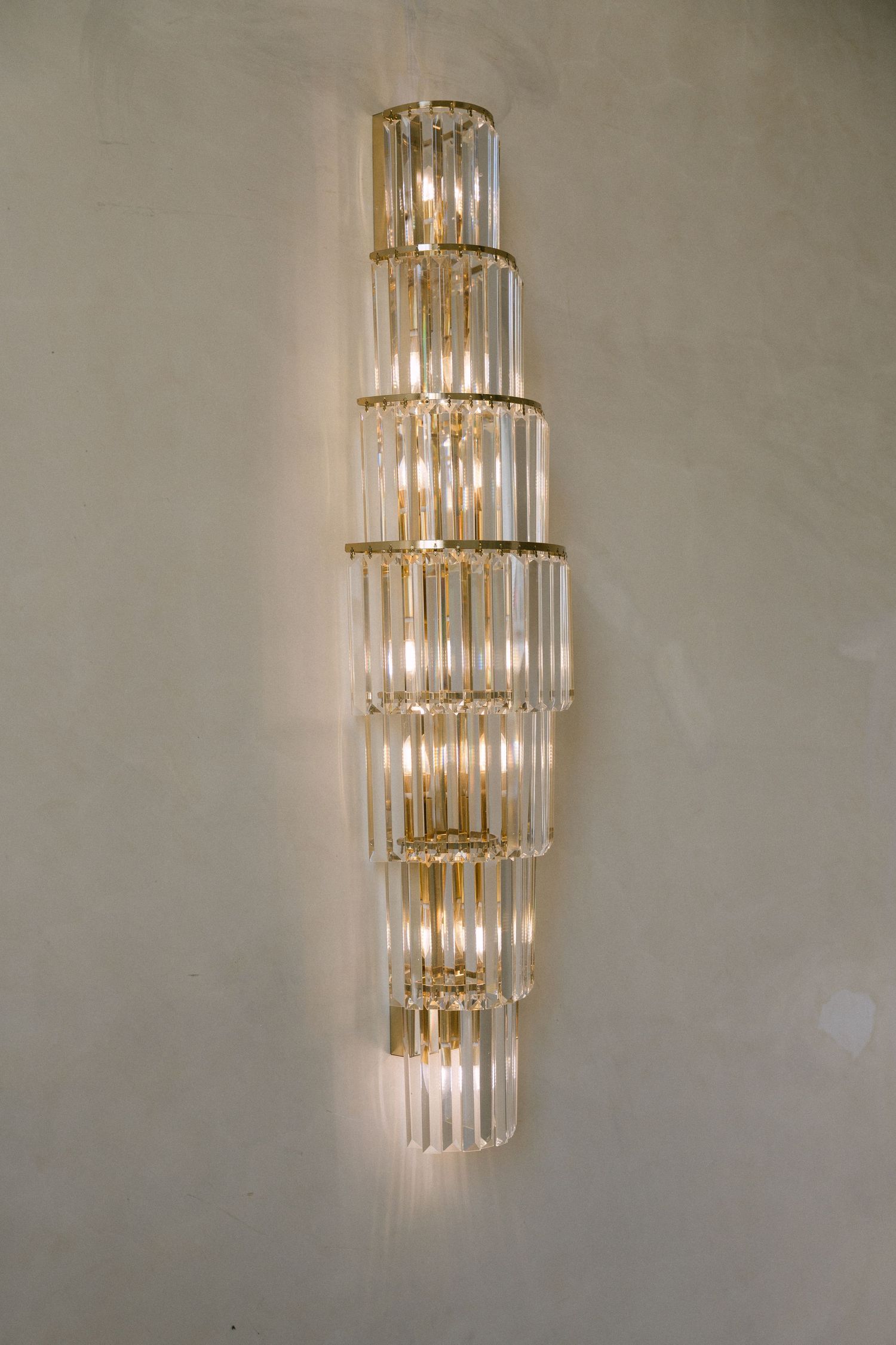 Elegant crystal wall sconce with gold frame featuring tiered glass rods illuminated against a neutral wall.