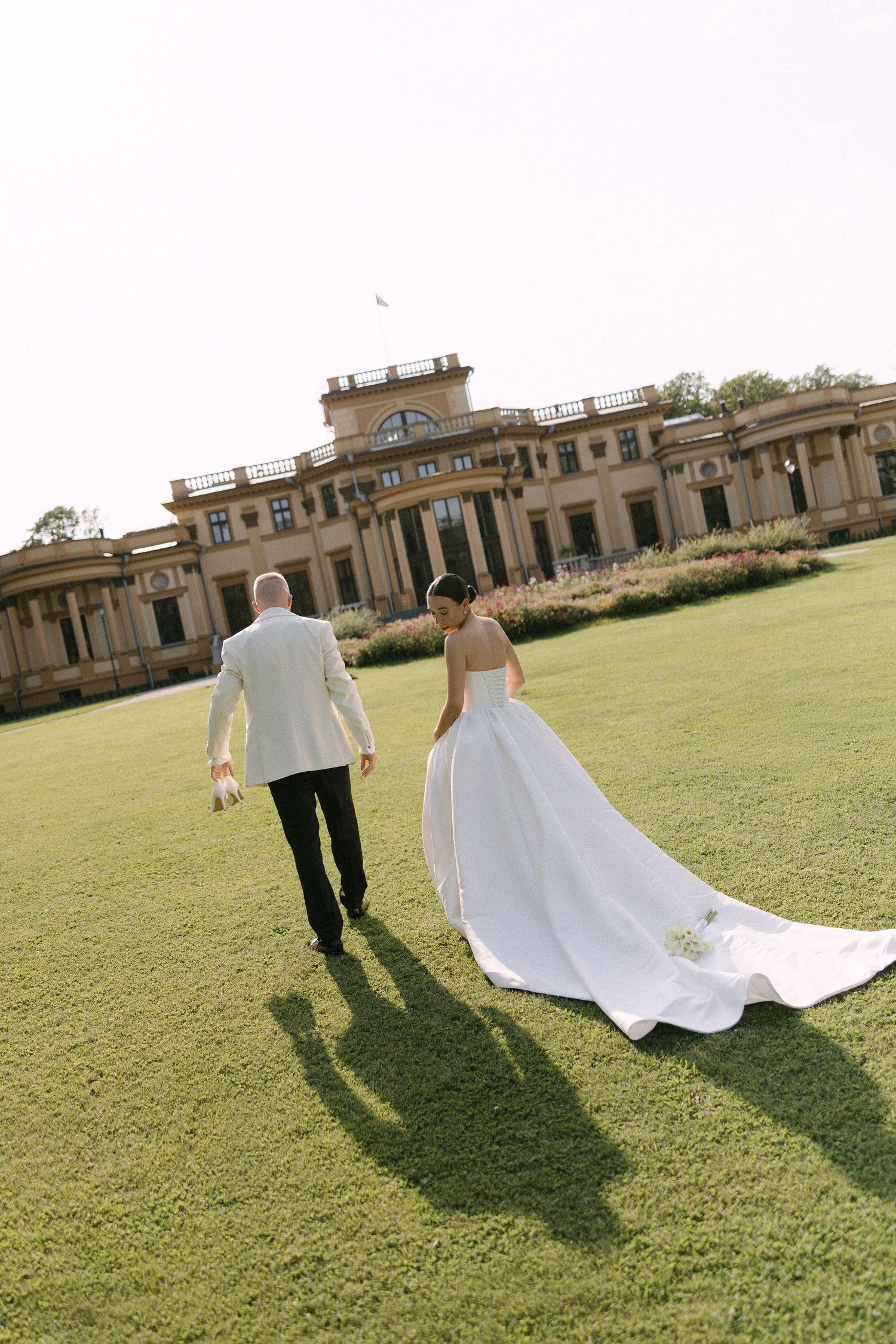 Bride and groom walking hand in hand across green lawn toward elegant estate venue on their wedding day.