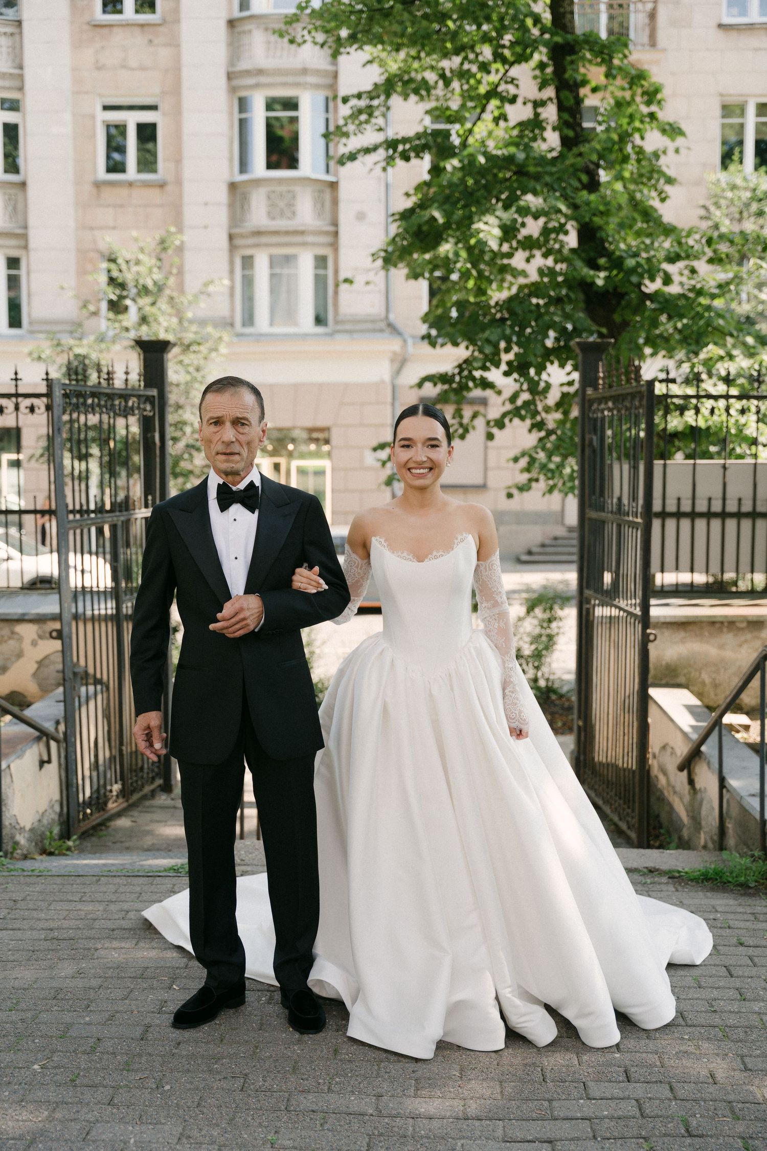Smiling bride in white ballgown with lace sleeves walks with groom in black tuxedo outdoors.