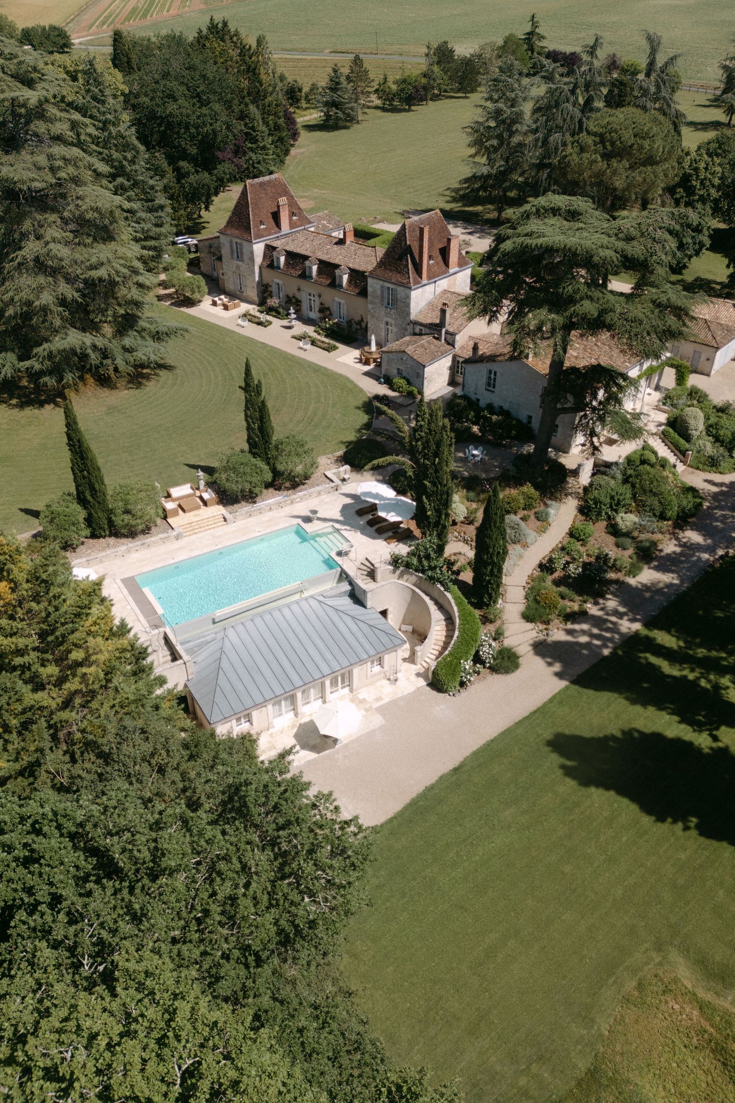 Aerial view of a French château with a swimming pool, manicured lawns, and surrounding woodland on a sunny day.