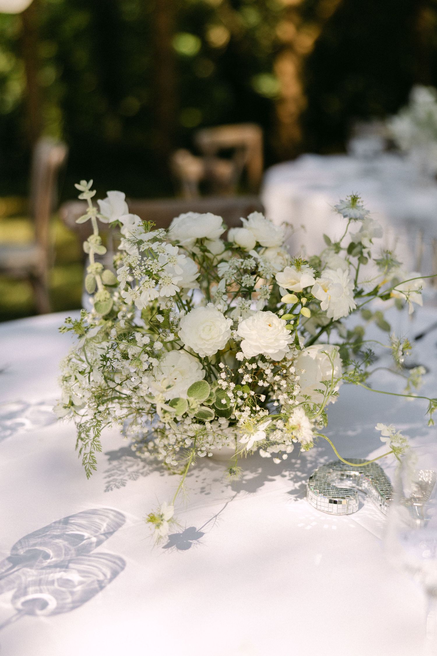 Elegant white floral centerpiece with roses and greenery on white linen reception table.
