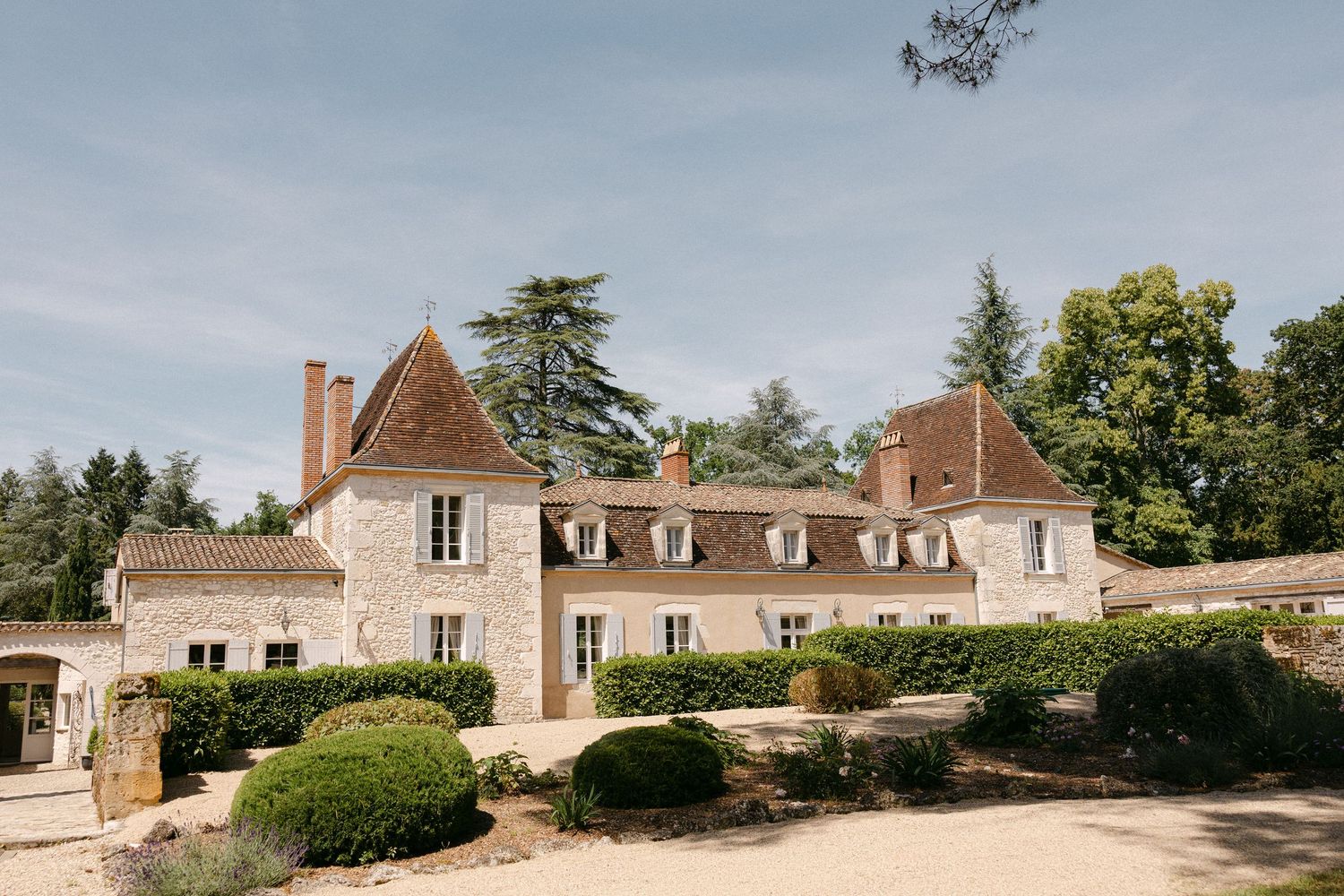 A charming French countryside manor house with stone walls, terracotta roof, and manicured hedges under a blue sky.