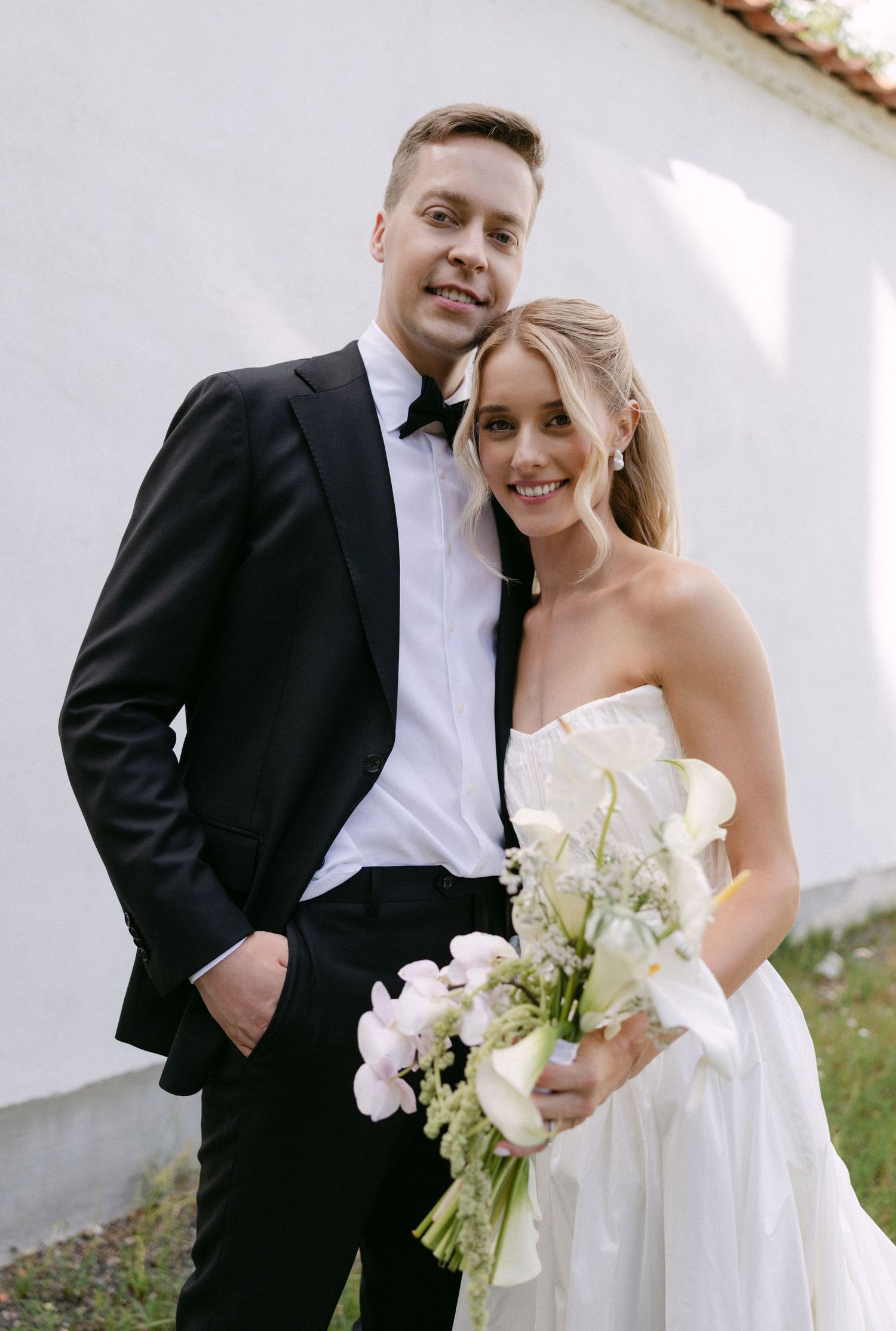 Bride and groom pose together smiling against a white wall, bride holding a white floral bouquet.