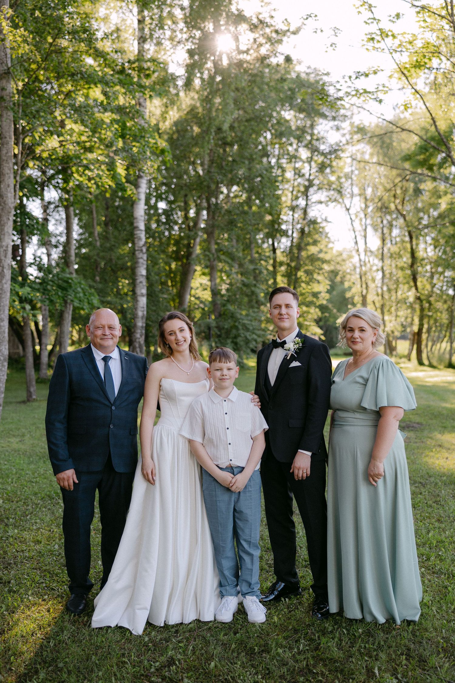 Wedding party of five posing outdoors on green grass, bride in white gown with groom in tuxedo and family members smiling.