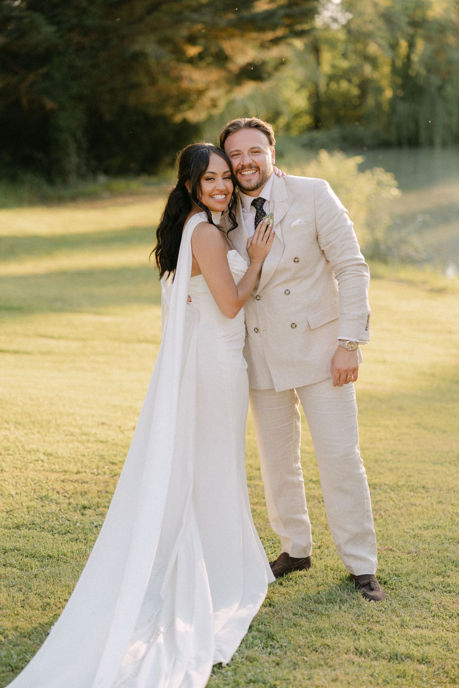 Smiling bride in white cape gown and groom in beige suit embrace on a golden sunlit lawn.
