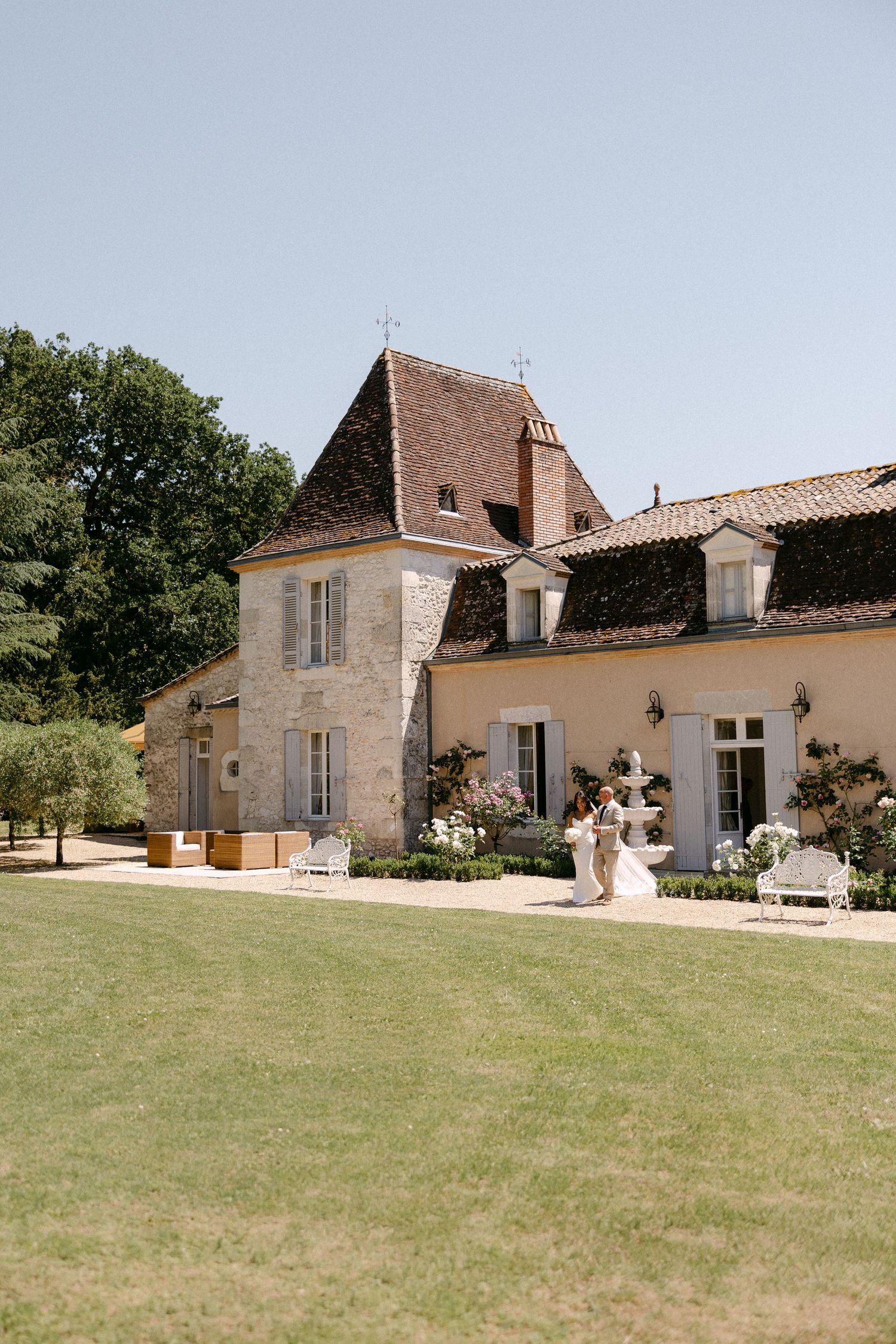 Elegant French chateau wedding venue with manicured lawn and stone tower under clear blue sky.