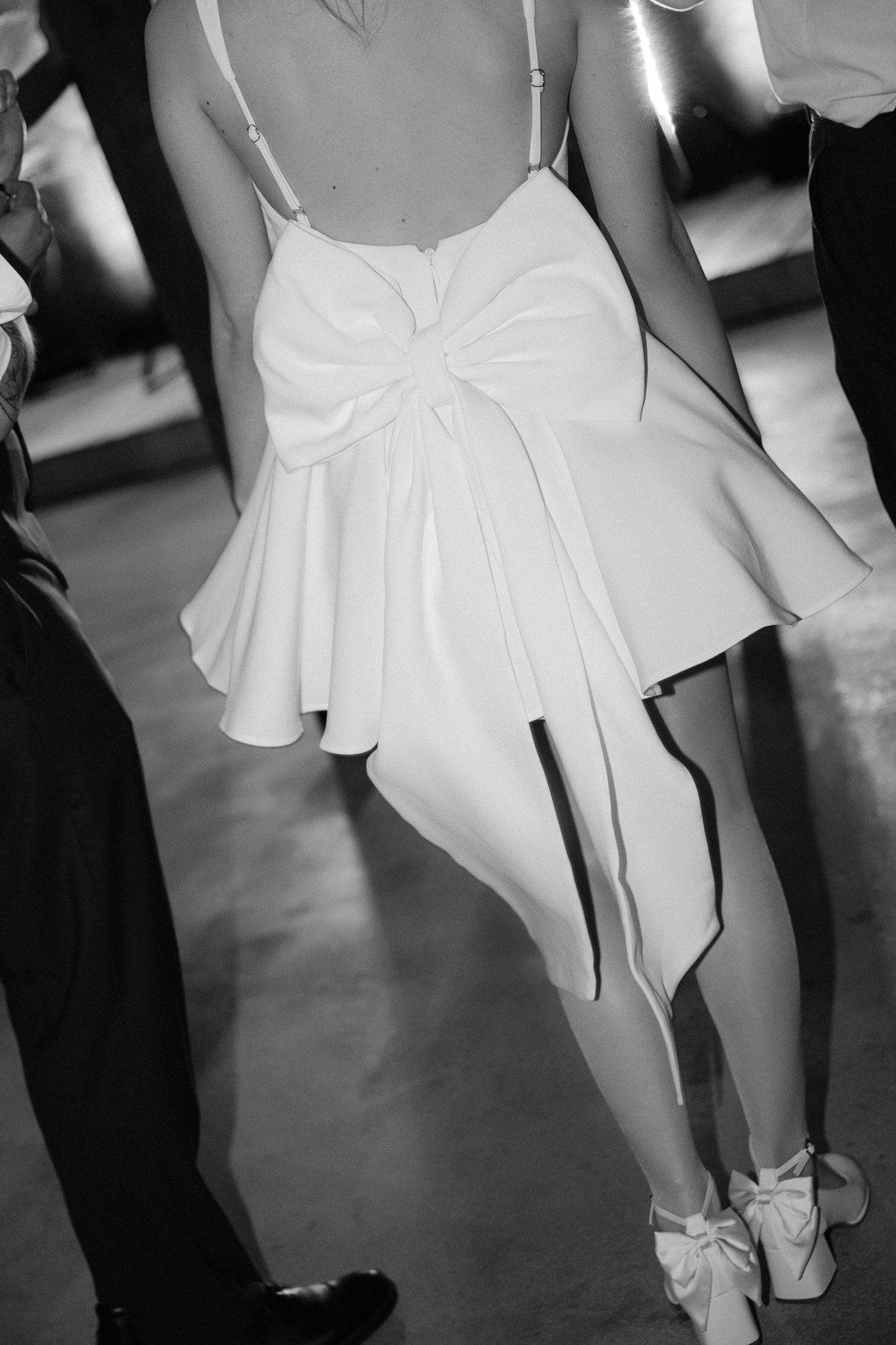 Black and white photo of bride in short white bow-back dress and heels dancing at wedding reception.