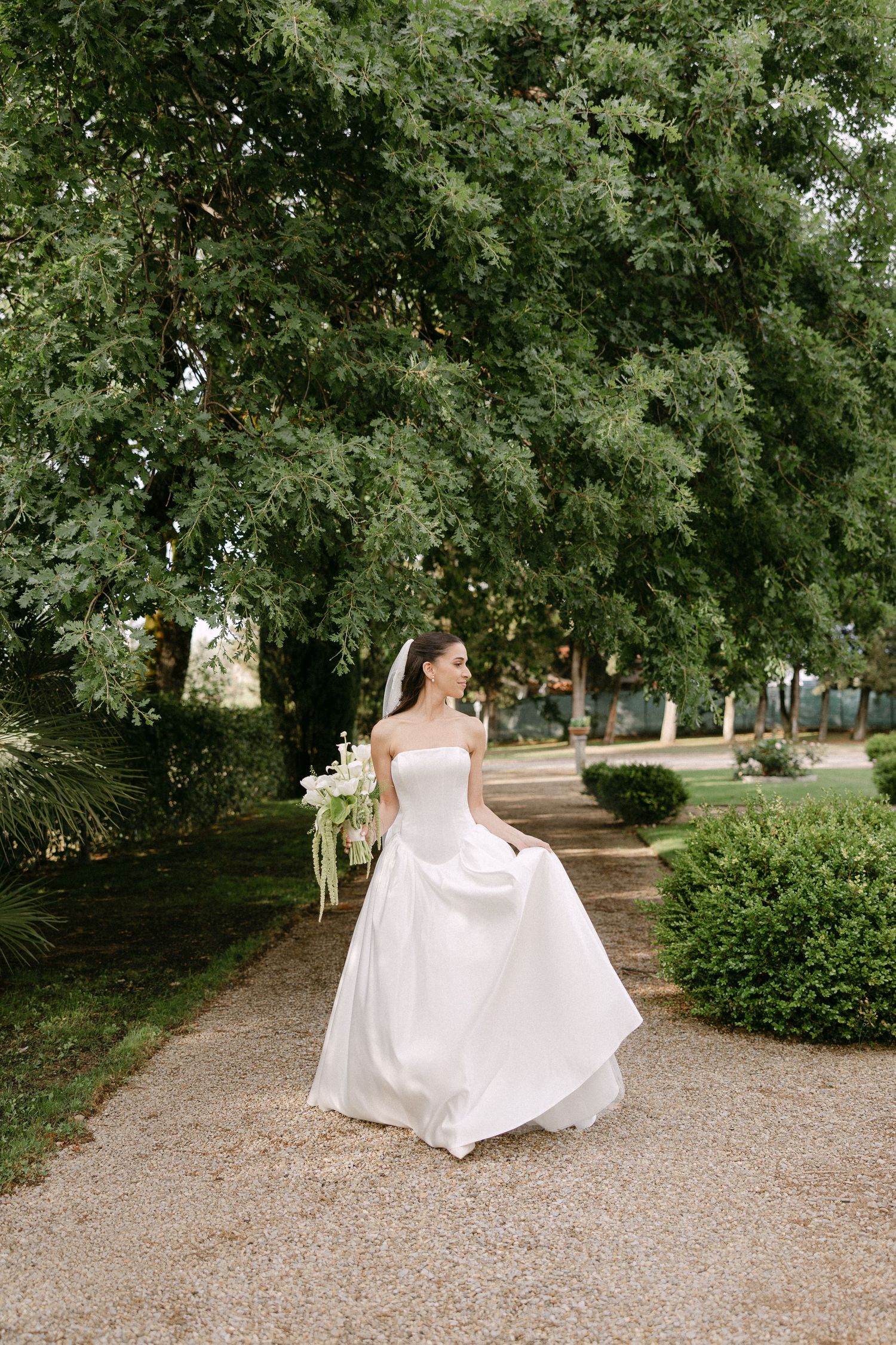 Elegant bride in strapless white ball gown walks along gravel garden path beneath lush green trees.