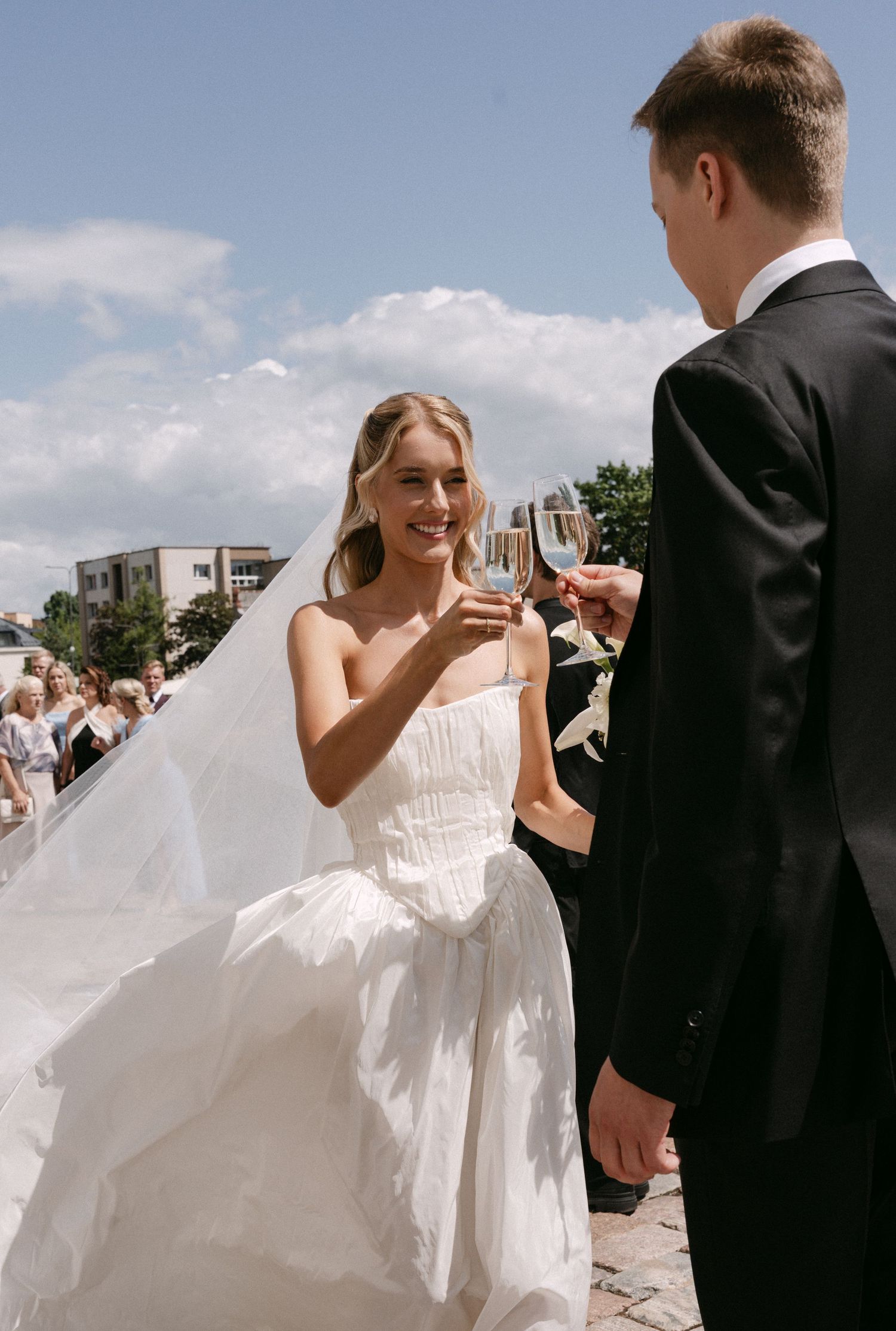 Smiling bride in white dress toasts champagne with groom in black suit outdoors on wedding day.