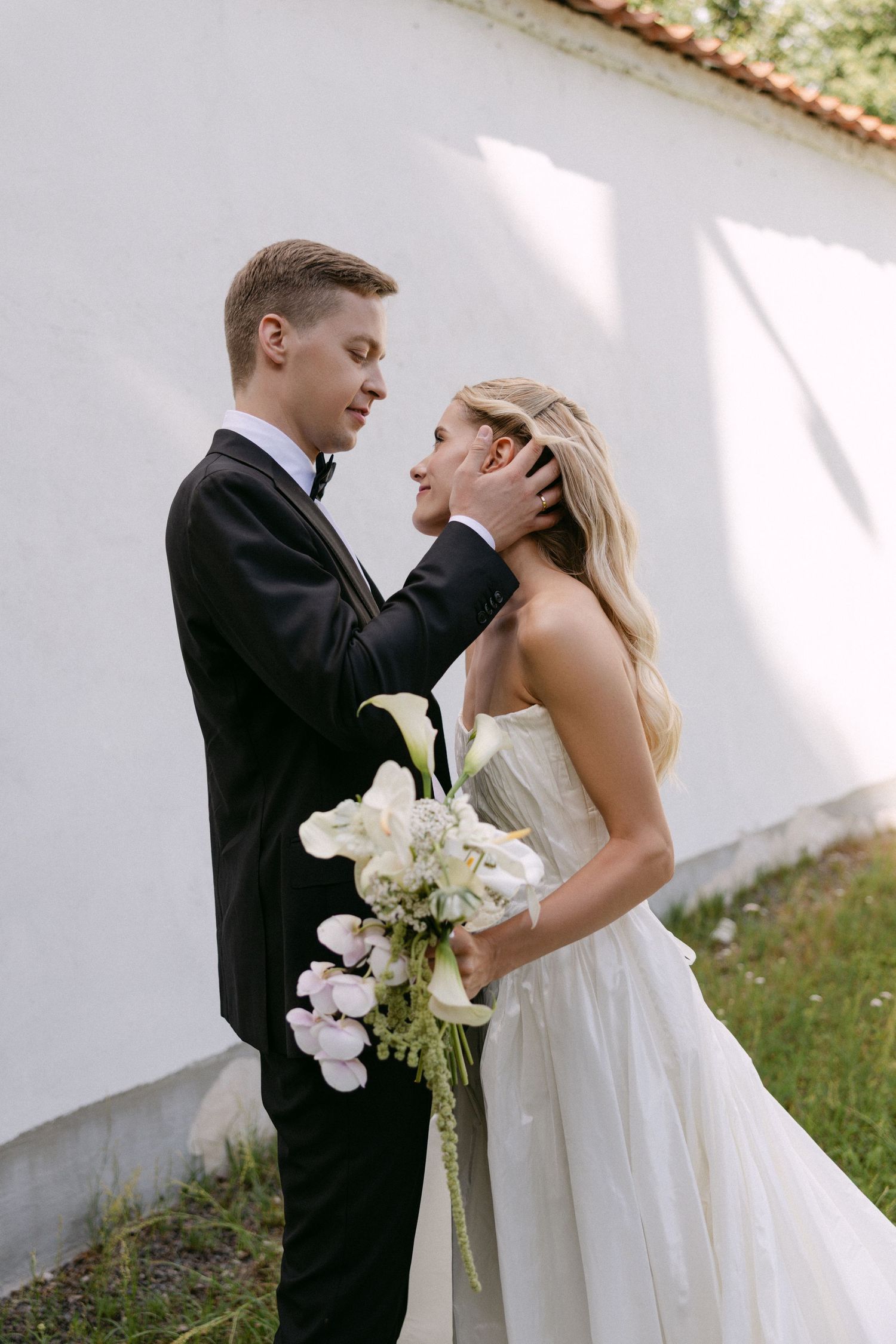 Groom in black tuxedo tenderly holding bride's face while she holds white calla lily bouquet.