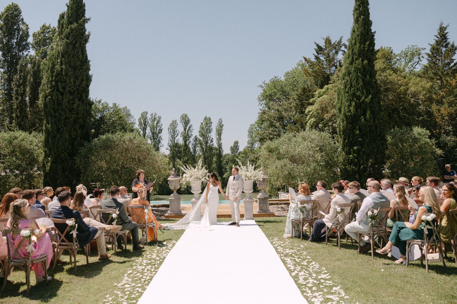 Outdoor wedding ceremony with white aisle, tall cypress trees, and seated guests on sunny day.